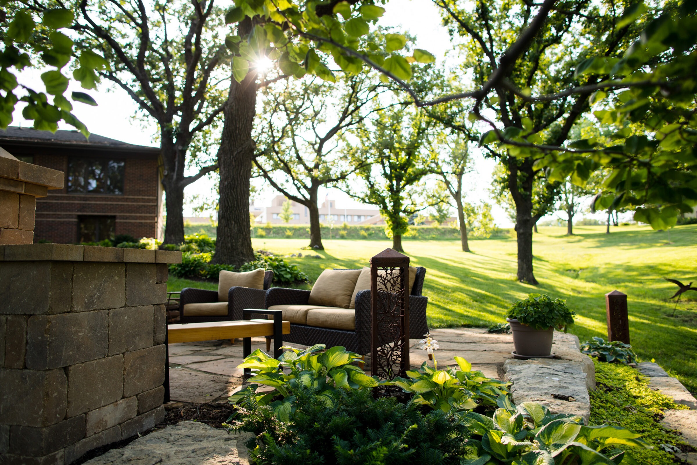 A peaceful patio with comfortable seating, surrounded by green plants and trees, under gentle sunlight. A house is visible in the background.