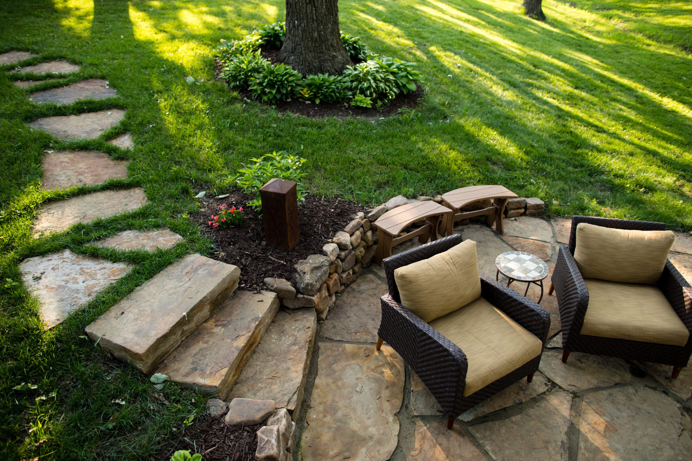 Cozy outdoor patio with wicker chairs, a small round table, stone pathway, surrounded by lush green grass and plants under tree shade.