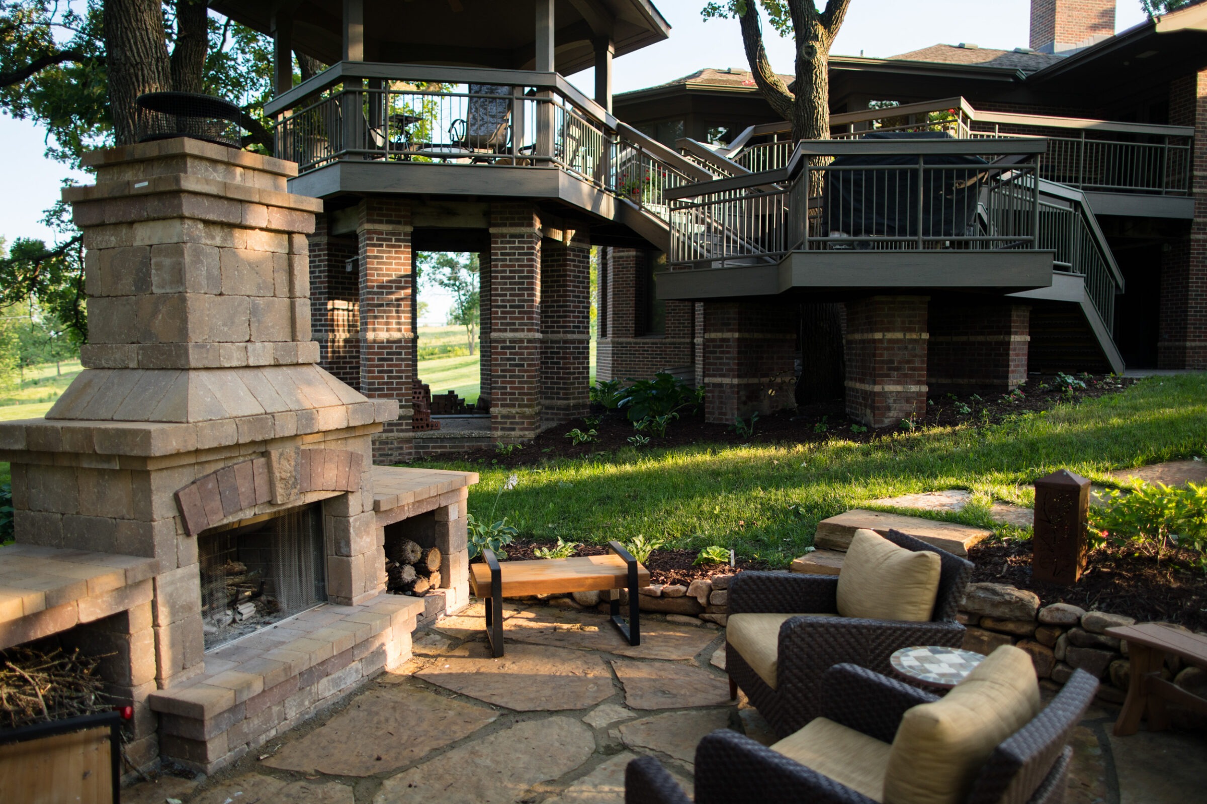 Patio with wicker furniture, stone fireplace, and elevated deck, surrounded by lush greenery. No recognizable landmarks or historical buildings are visible.