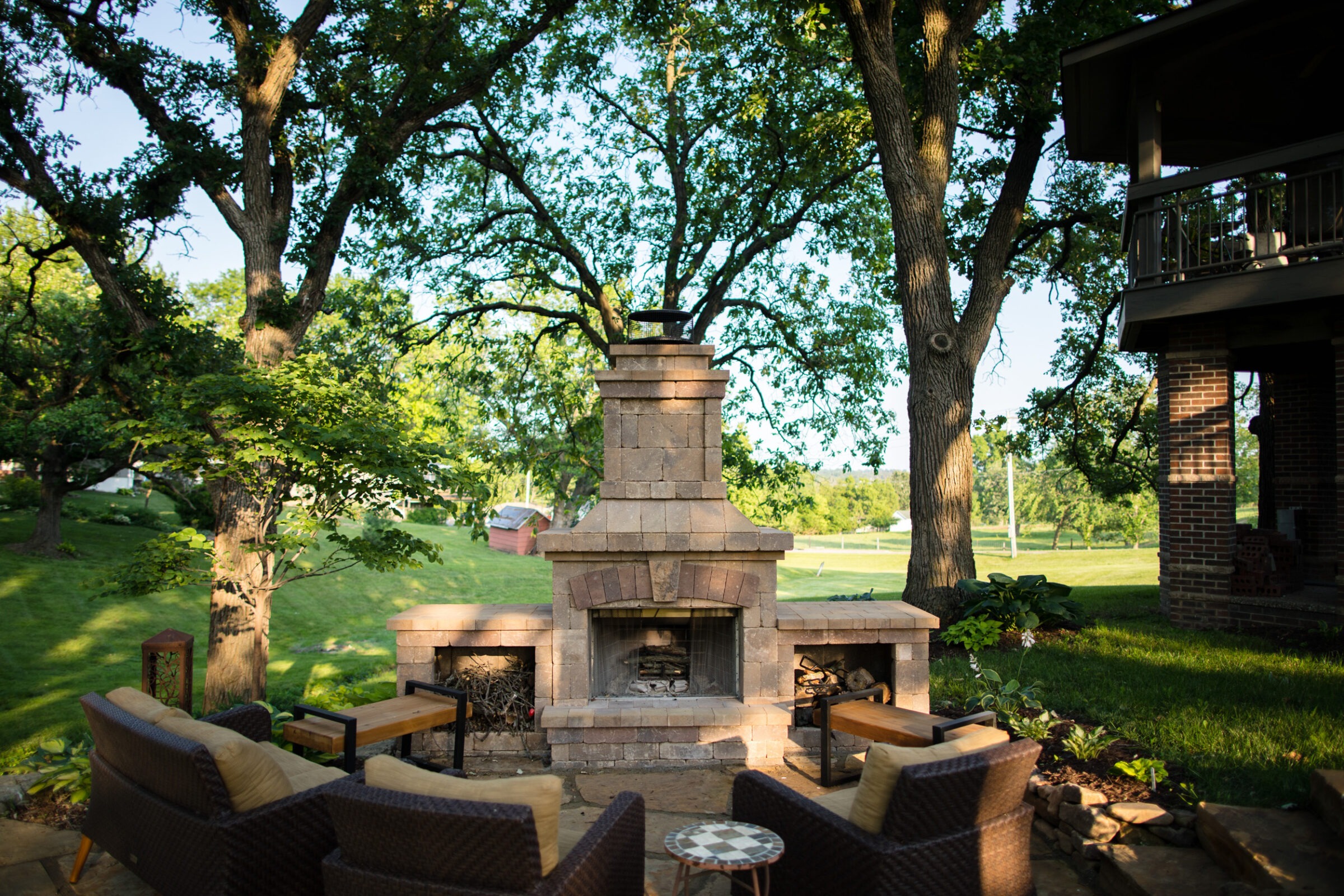 Outdoor patio featuring a stone fireplace, surrounded by wicker furniture and lush greenery, shaded by large trees, creating a relaxing atmosphere.