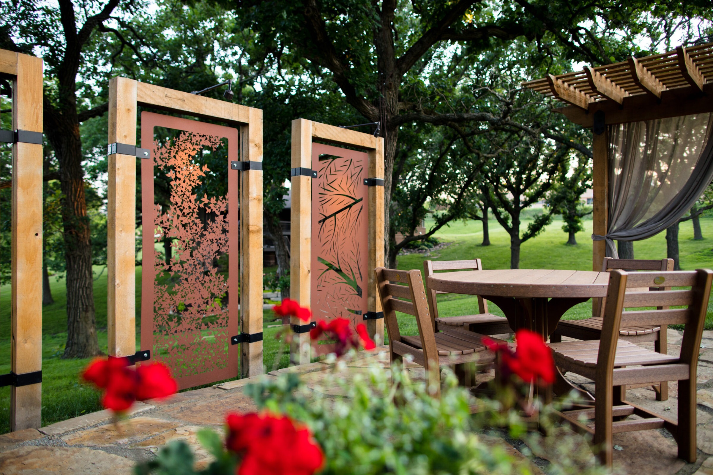 Wooden outdoor dining area with decorative metal panels, pergola, and red flowers. Lush green trees surround the peaceful garden setting.