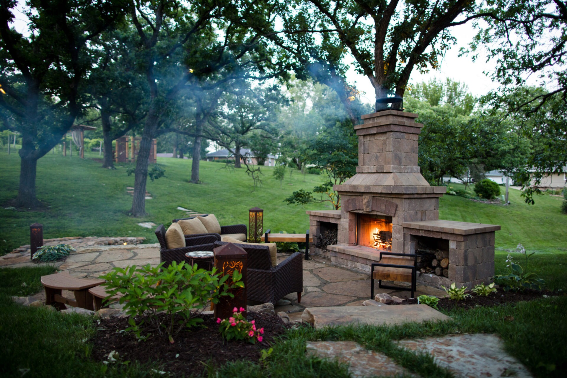 Outdoor seating area with a stone fireplace, surrounded by grassy lawn and trees. Cozy atmosphere with wicker chairs and vibrant plants.