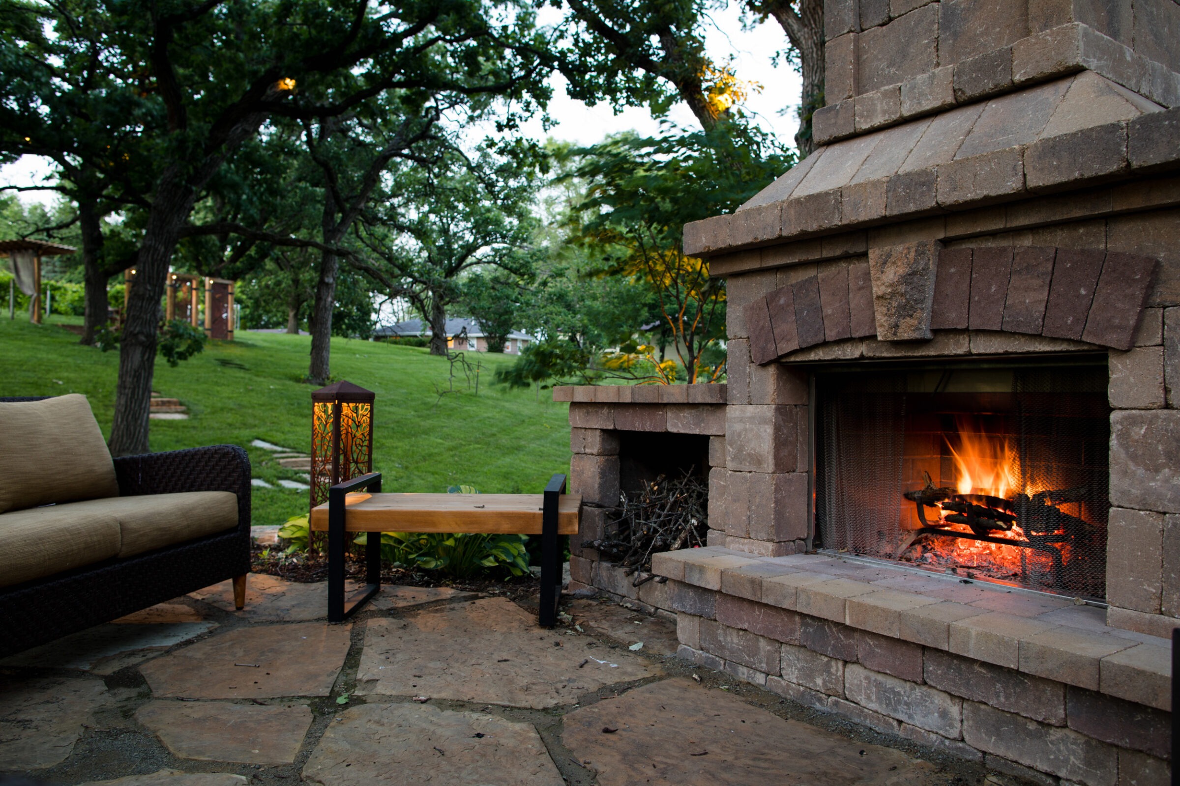 Cozy patio features stone fireplace with a blazing fire, surrounded by seating, lush green trees, and ambient lantern lighting in tranquil garden.