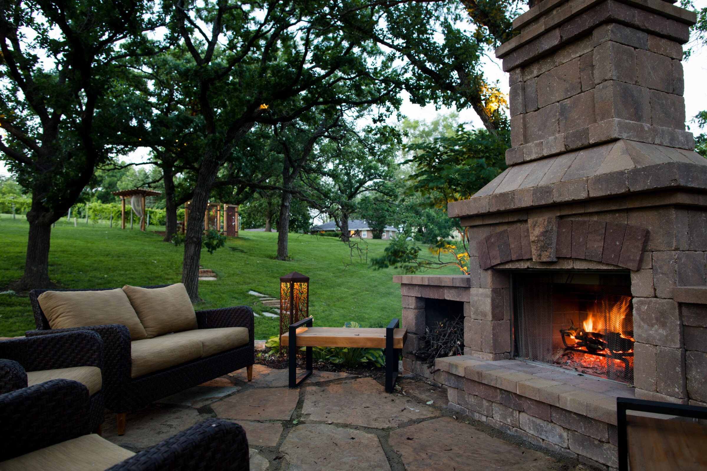 Outdoor patio with a lit fireplace, cushioned seating, and lanterns. Surrounded by lush green trees and a grassy landscape.