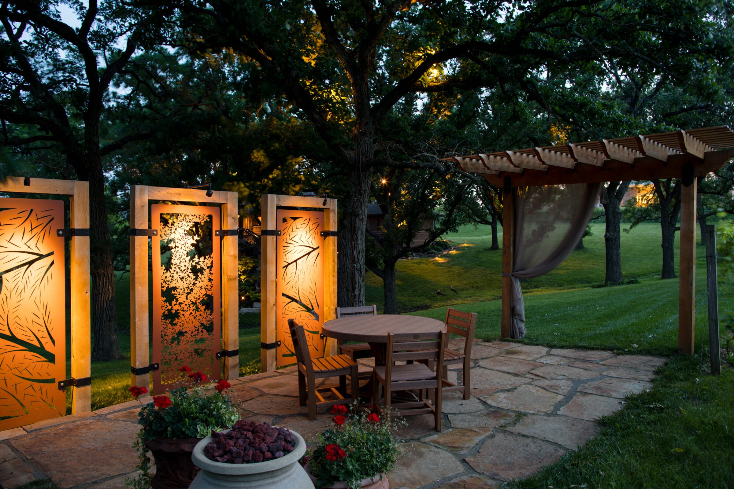 A serene garden patio at dusk, featuring illuminated decorative panels, wooden furniture, and a pergola surrounded by lush trees and greenery.