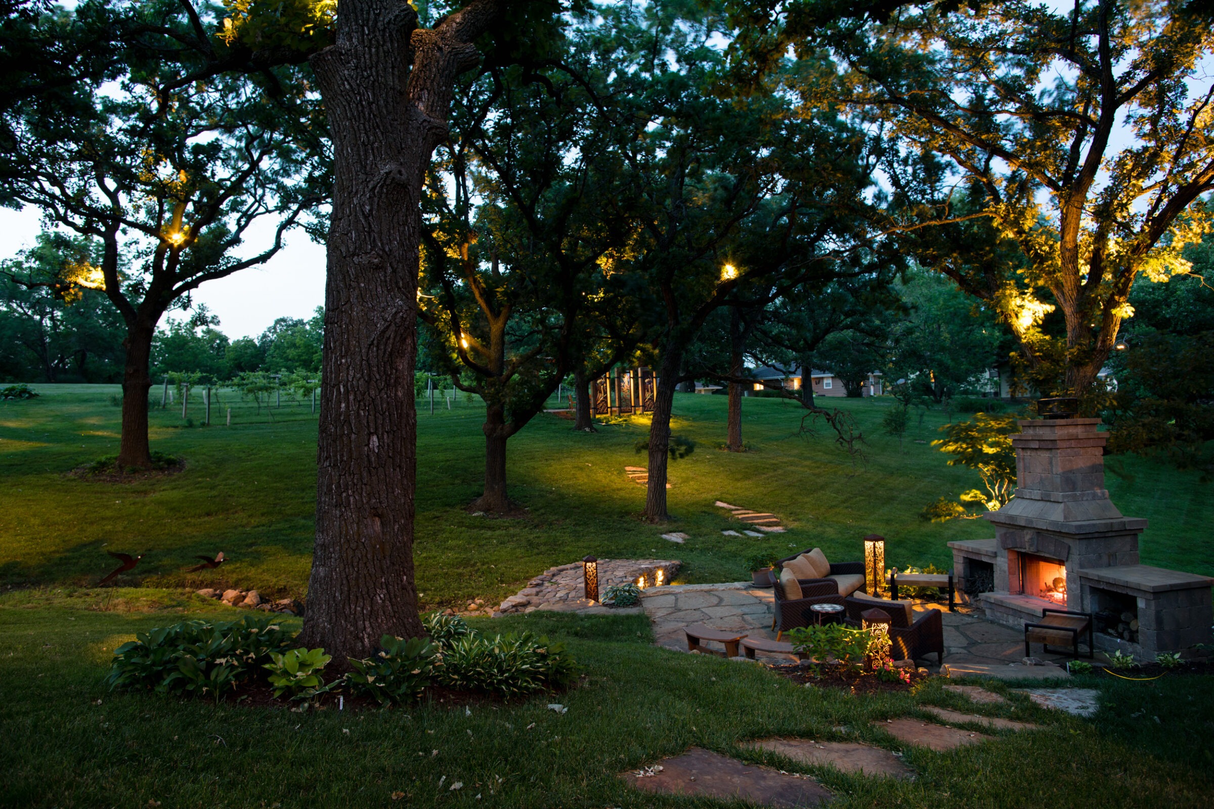 A cozy outdoor sitting area with a lit fireplace, surrounded by trees and garden lights creating a serene evening atmosphere.