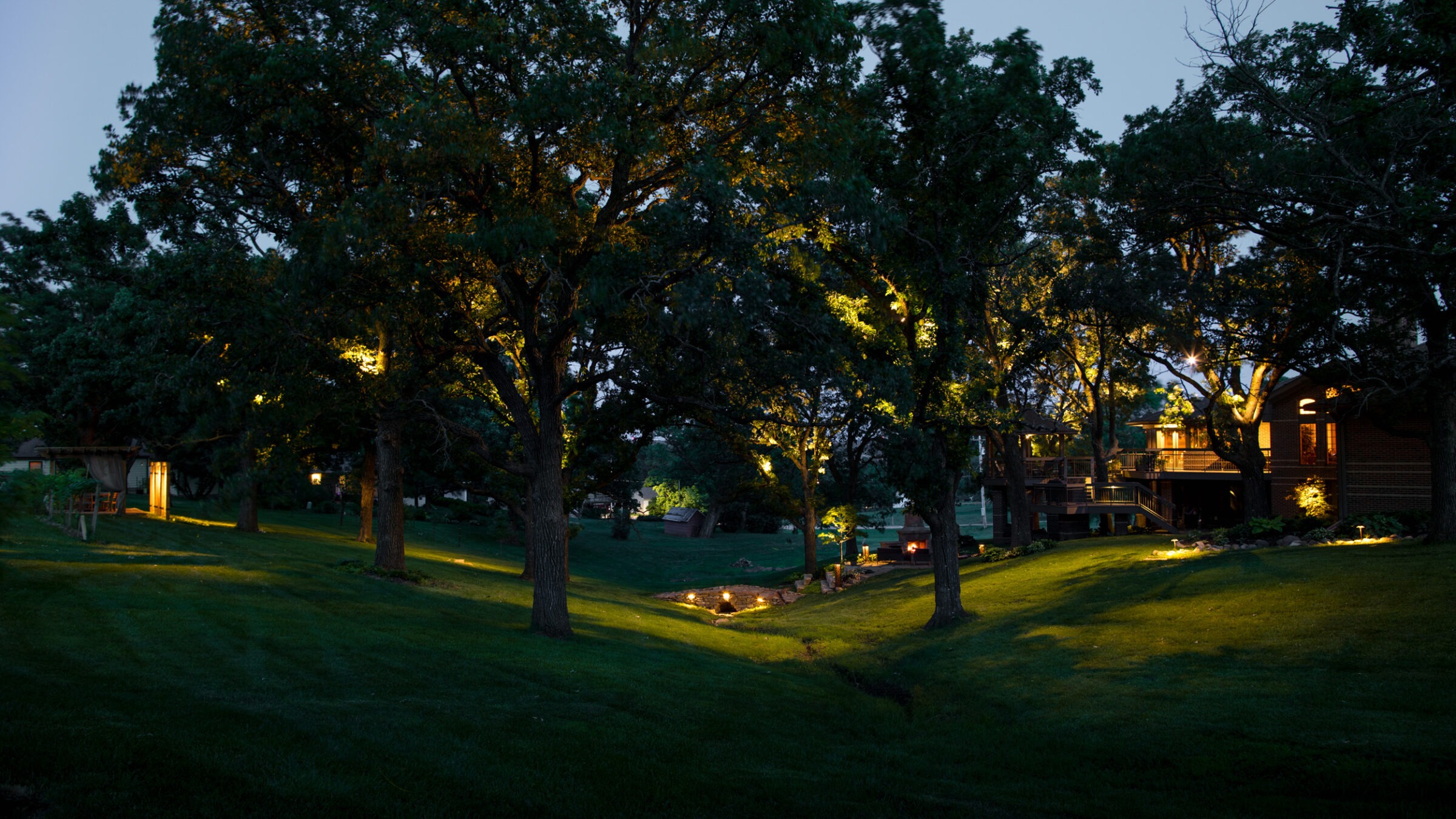 A serene, well-lit backyard garden with tall trees, glowing pathways, and a house nestled among vibrant greenery at dusk.