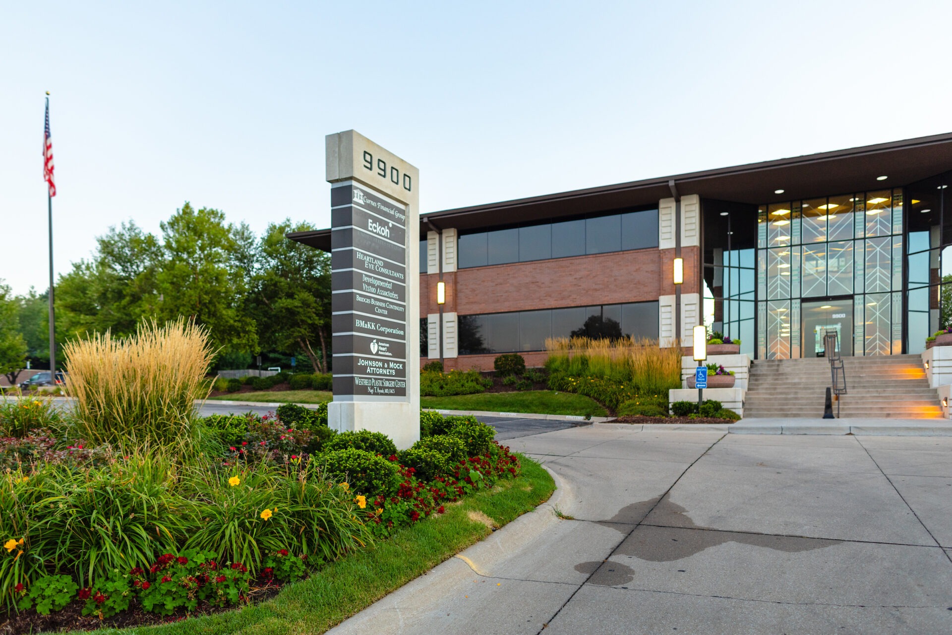 Modern office building with glass entrance, surrounded by landscaped gardens and a directional sign displaying business names. American flag visible in background.