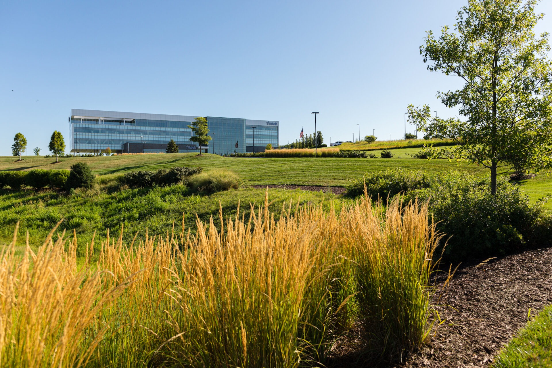 A modern glass building surrounded by green grass and trees under a clear blue sky. American flag visible in the background.