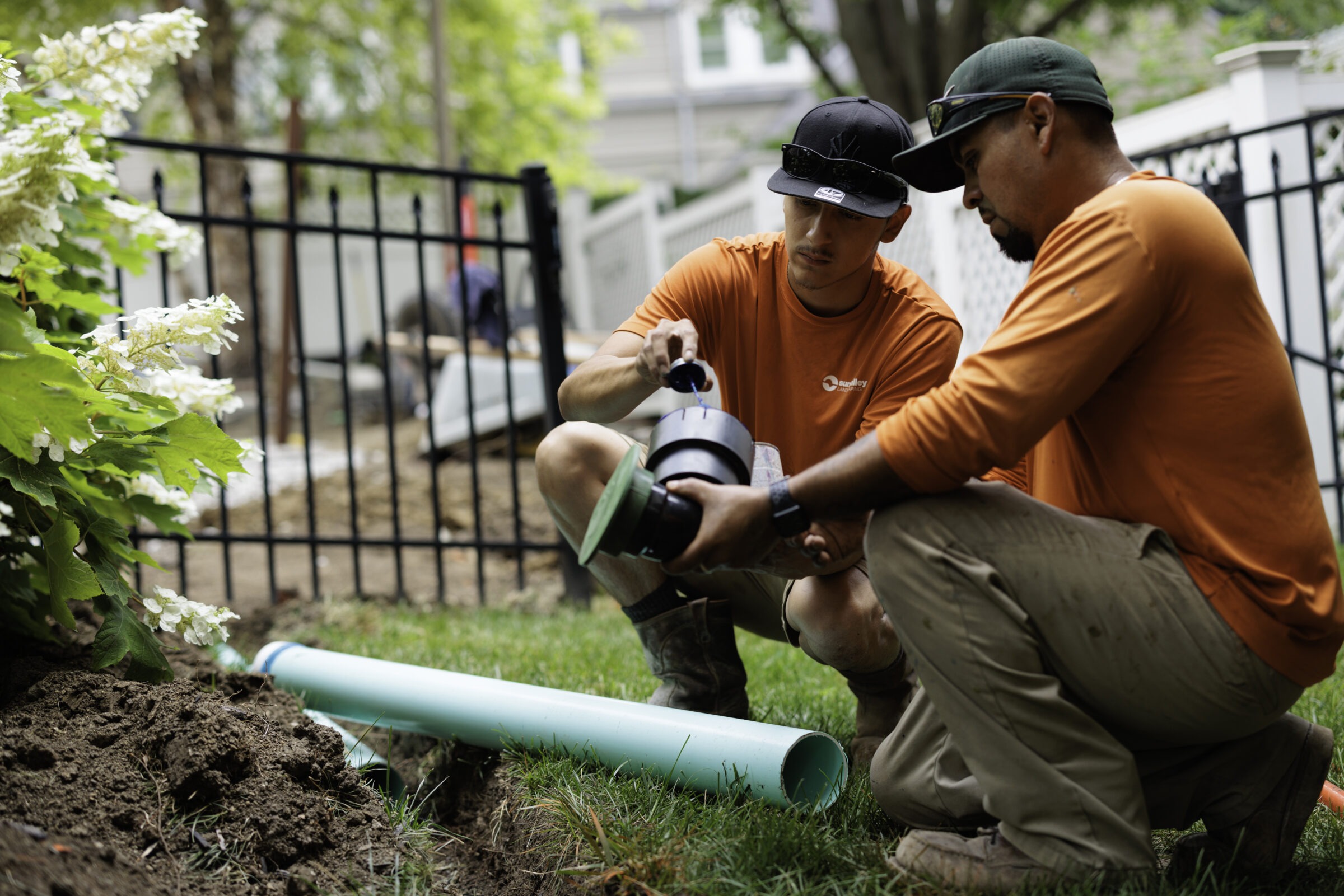 Two people work on a garden pipe installation, wearing orange shirts and hats, surrounded by greenery and a black metal fence.