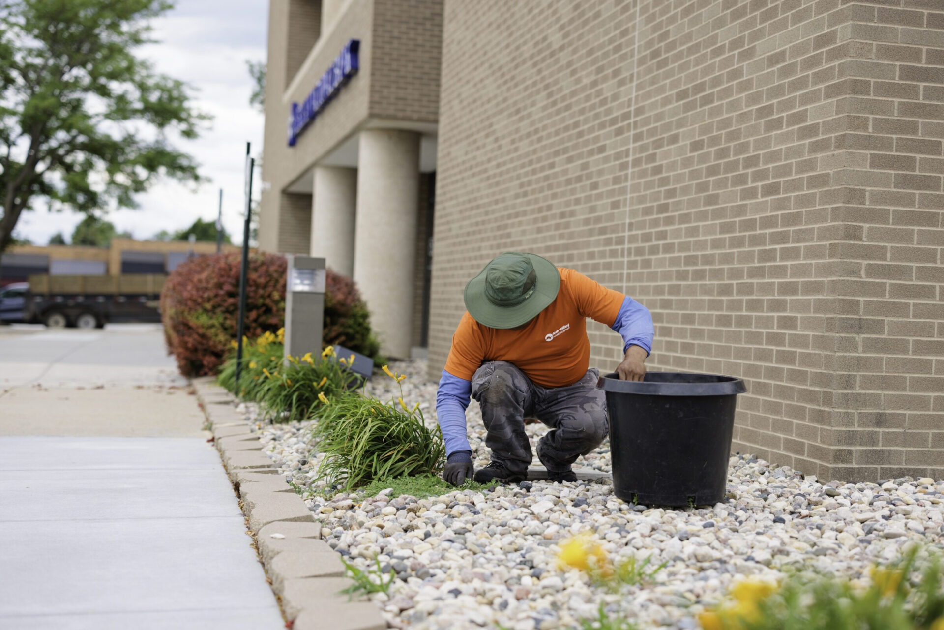 A person in an orange shirt tends a landscaped area with rocks and plants near a brick building under a cloudy sky.