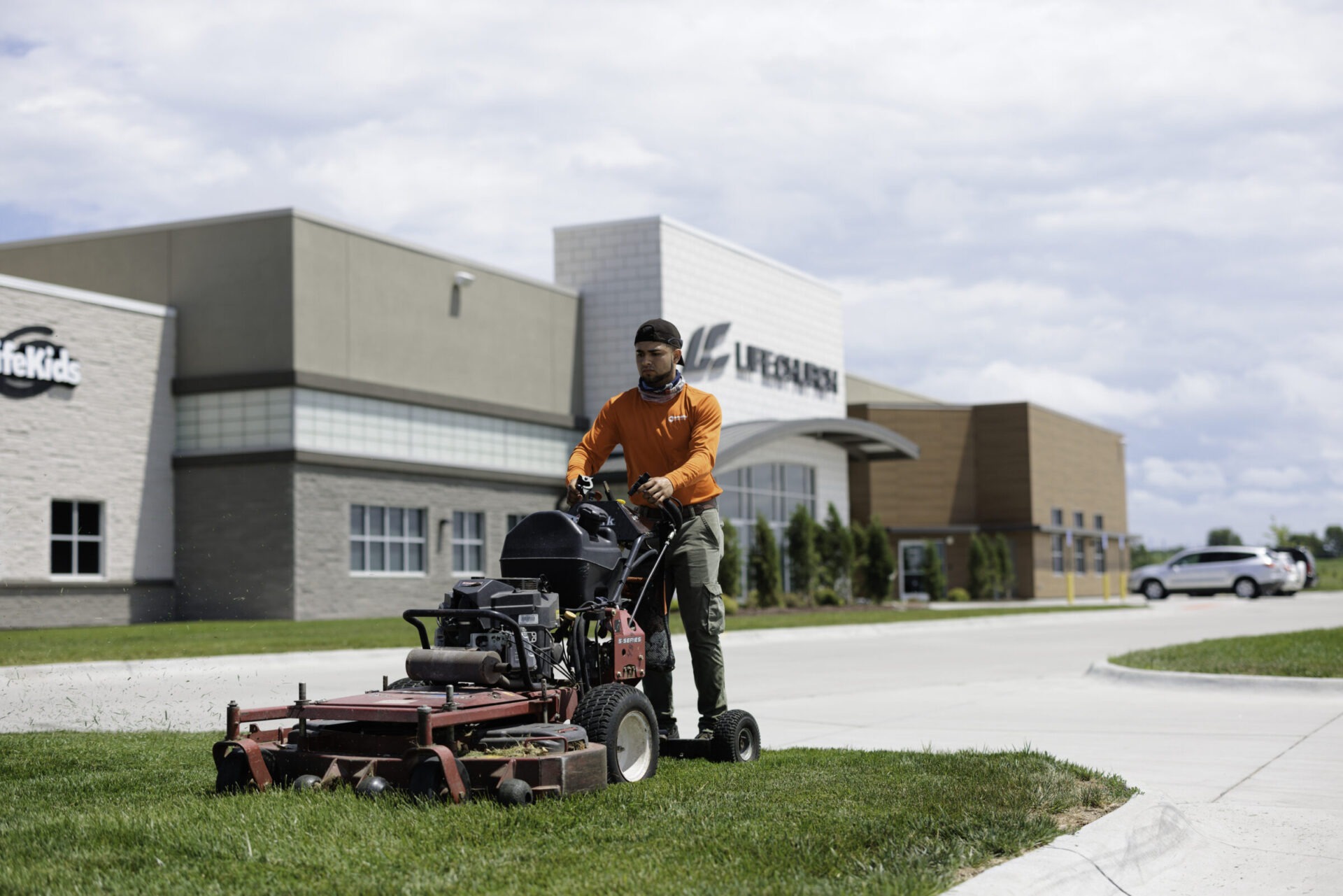 A person operates a lawnmower outside a modern building with "LifeCare" signage, under a cloudy sky. Nearby, a vehicle is parked.