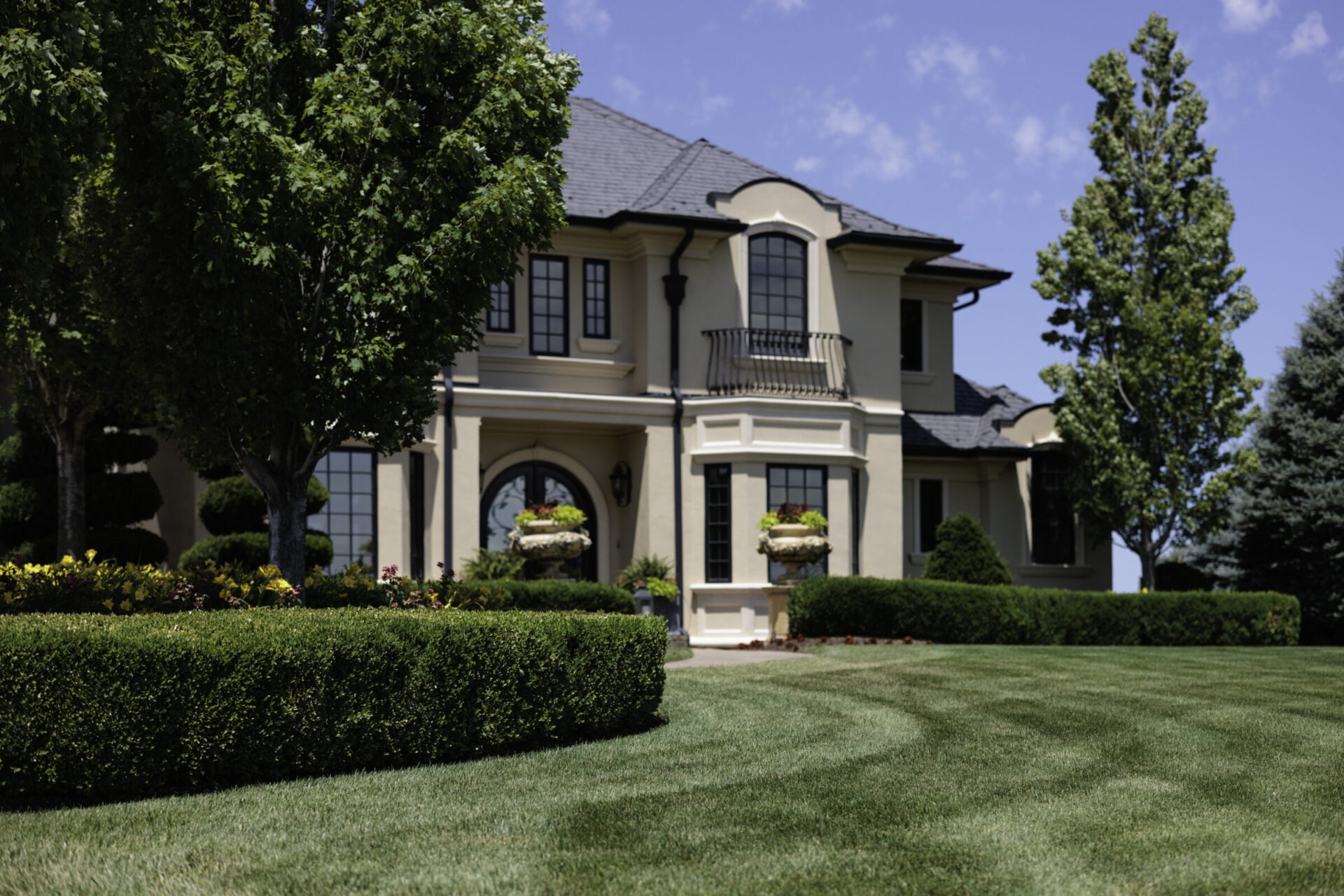 A luxurious house with manicured lawn, surrounded by trees and shrubs, under a clear blue sky on a sunny day.