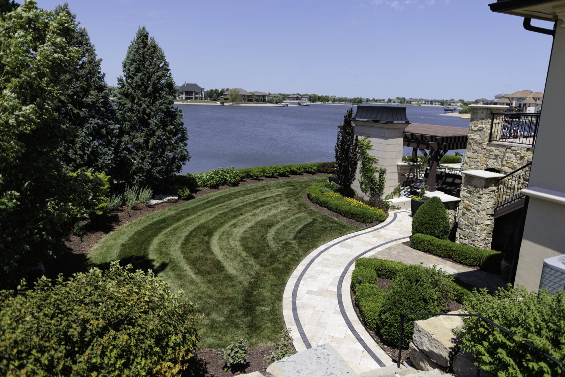 Beautiful landscaped garden with stone paths, lush greenery, and a lake view; residential homes line the opposite shore under a clear blue sky.