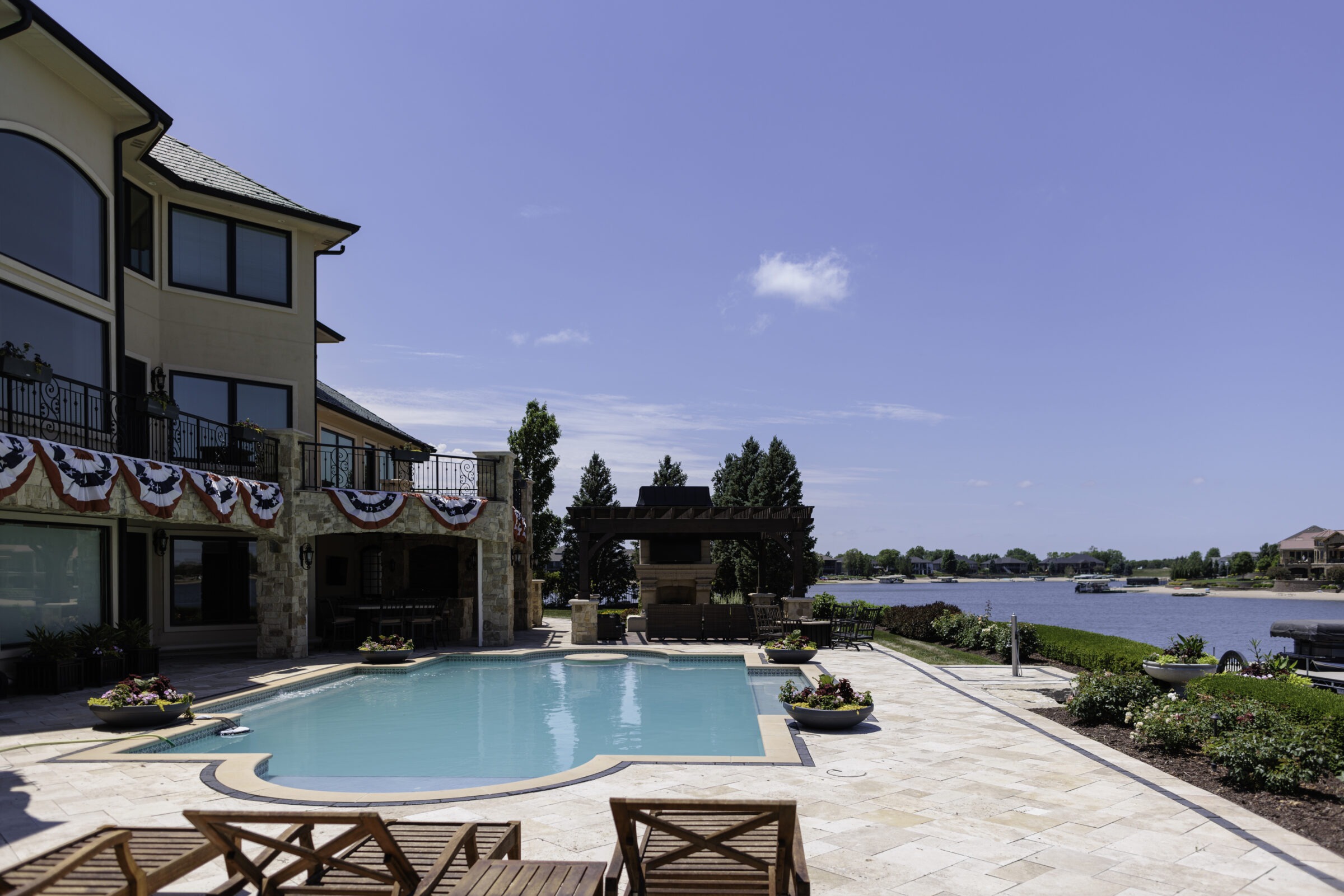 Luxurious lakeside house with a decorated terrace, swimming pool, and scenic water views under a clear, blue sky.