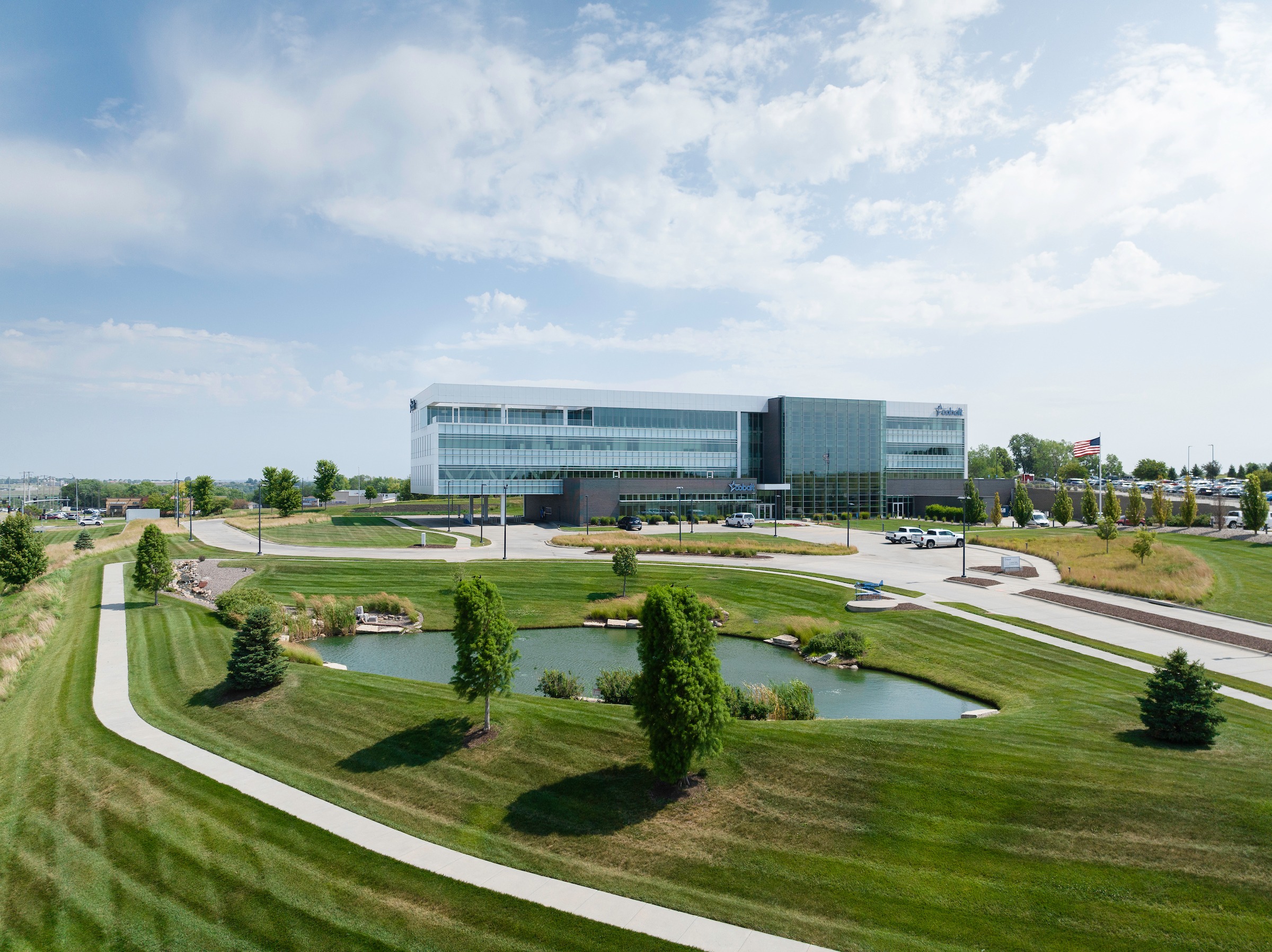 A modern glass building surrounded by lush lawns and a small pond under a clear sky, with a visible American flag nearby.