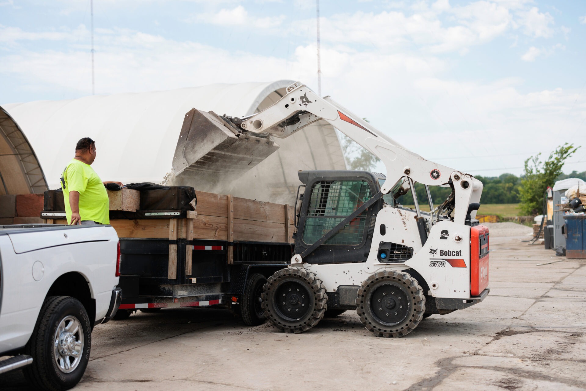 A person watches a Bobcat loader dumping materials into a truck on a construction site under a partly cloudy sky.