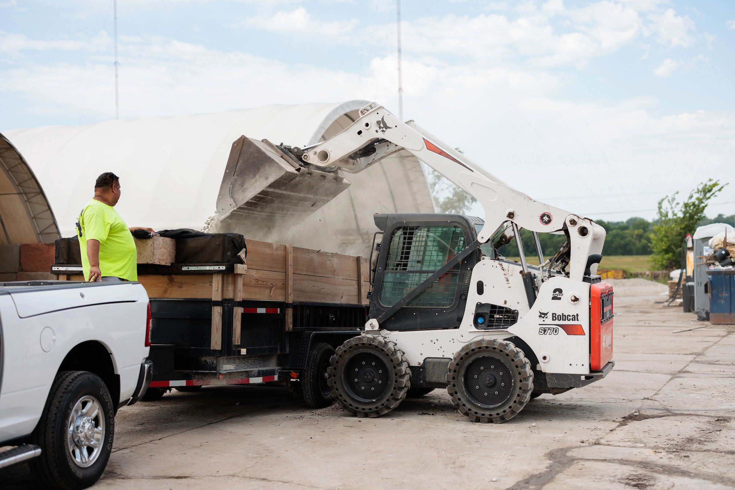 A person watches a Bobcat loader dumping materials into a truck on a construction site under a partly cloudy sky.