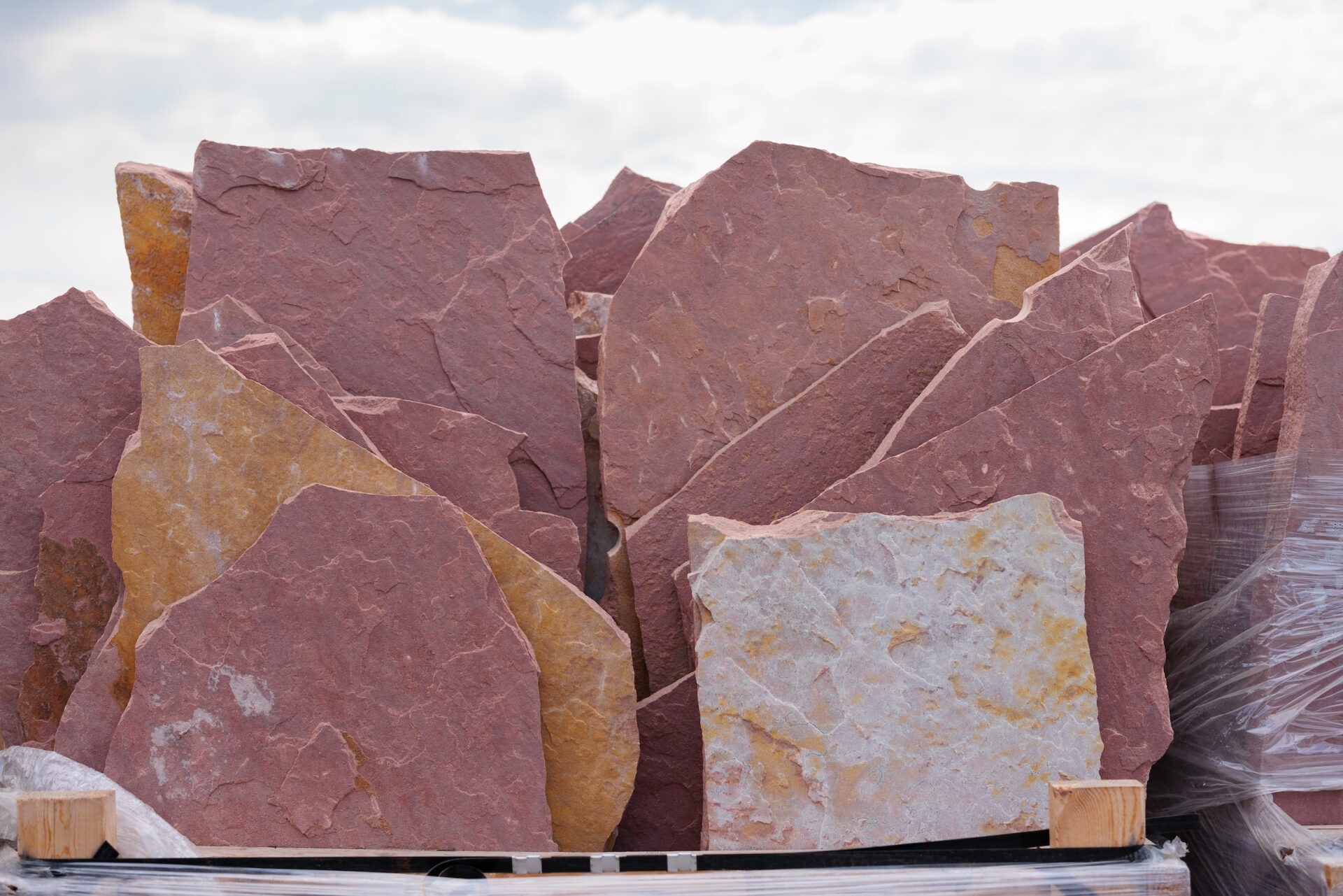 Large, flat pieces of reddish-brown sandstone stacked together under a cloudy sky, wrapped in plastic on a wooden pallet for transport.