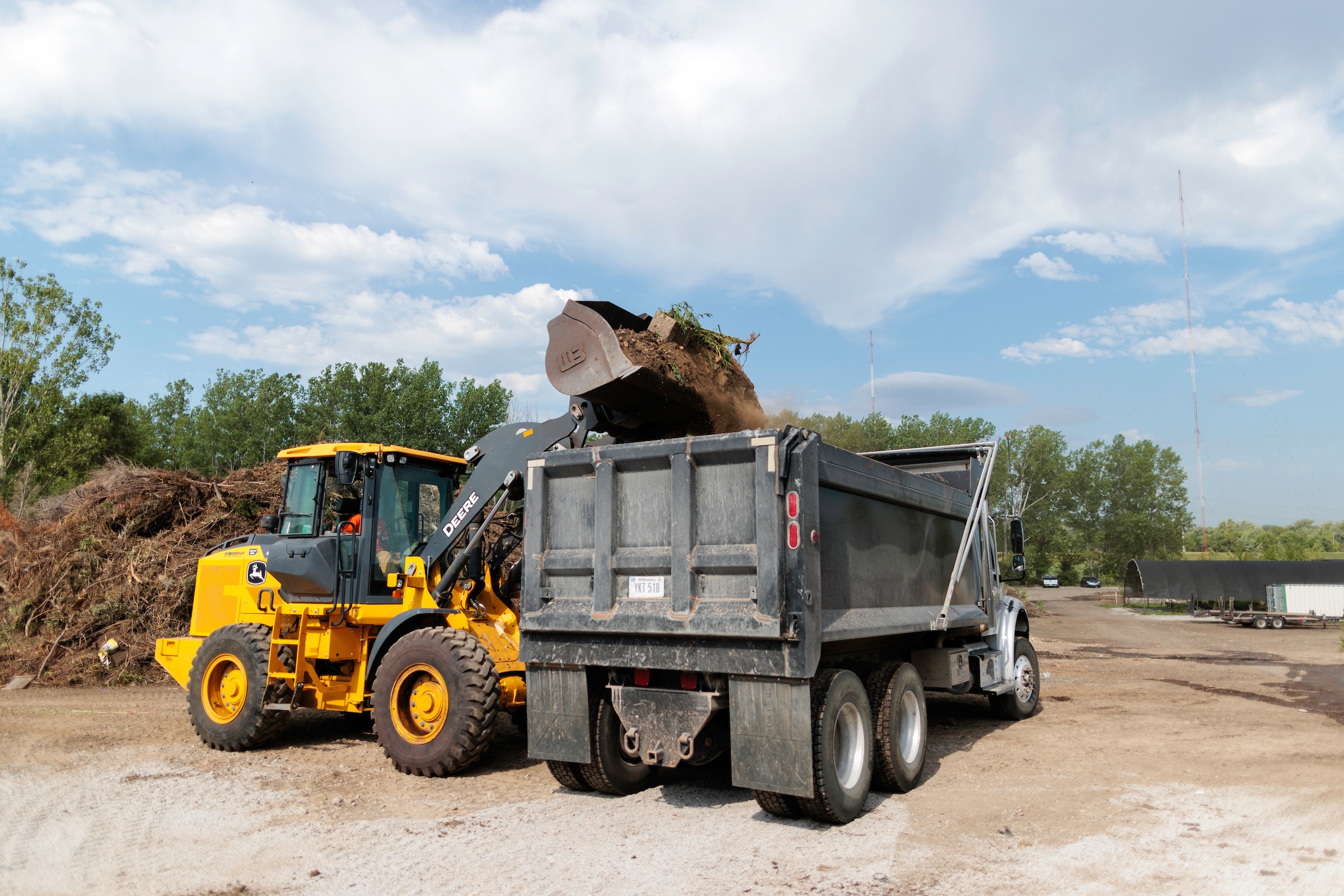 A yellow front-loader dumps soil into a large dump truck on a construction site surrounded by trees and clear skies.