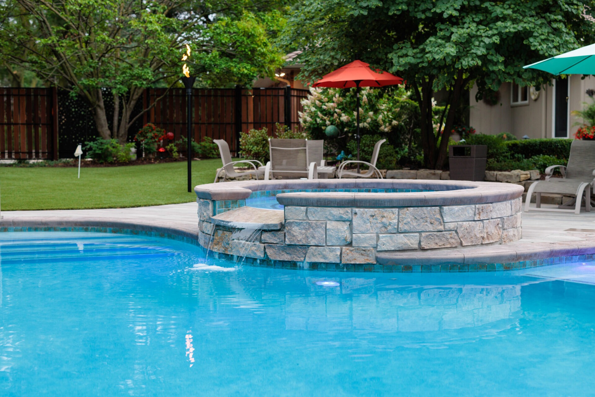 A serene backyard features a swimming pool with a stone spa, lounge chairs, and colorful umbrellas surrounded by lush greenery and trees.