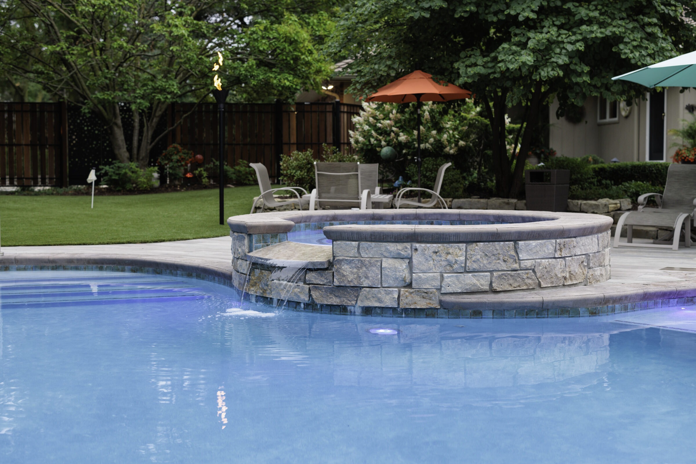 A serene backyard features a swimming pool, hot tub, lounge chairs, and umbrellas surrounded by lush greenery and a wooden fence.