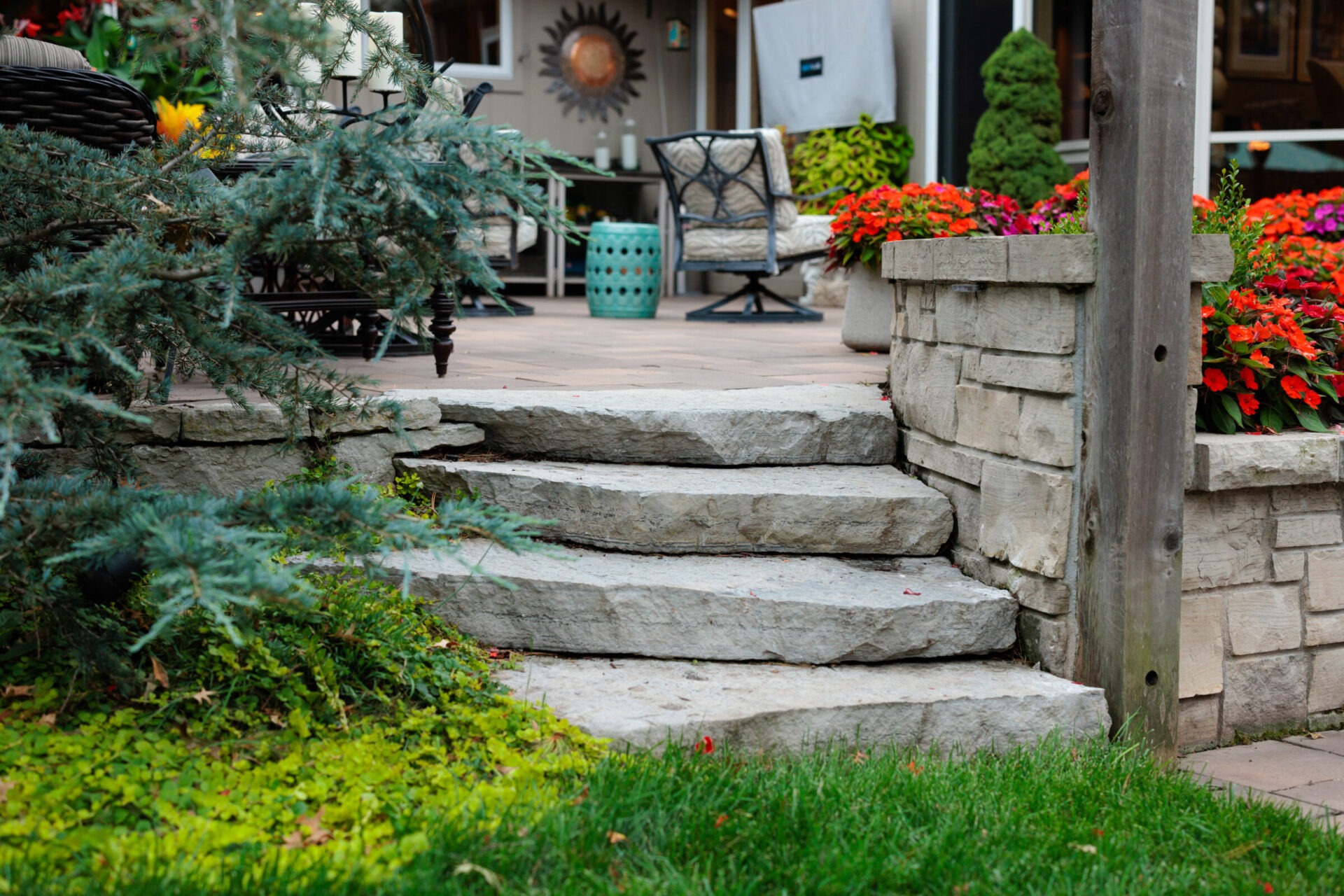 Stone steps lead to a patio with colorful flowers, outdoor furniture, tree branches, and green grass in a cozy garden setting.