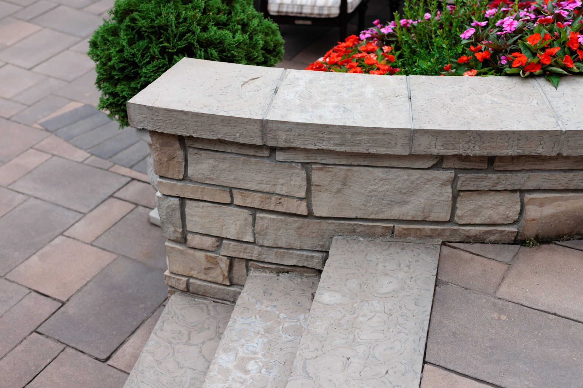 Stone steps next to a short stone wall, lush greenery, and colorful flowers on a paved patio area, creating a peaceful outdoor setting.