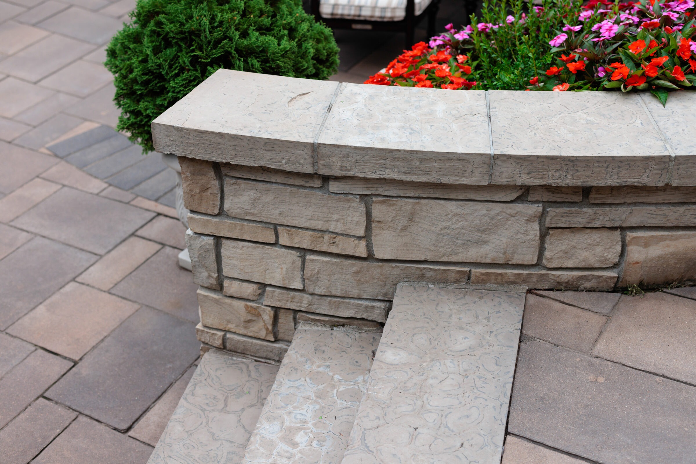 Stone steps next to a short stone wall, lush greenery, and colorful flowers on a paved patio area, creating a peaceful outdoor setting.