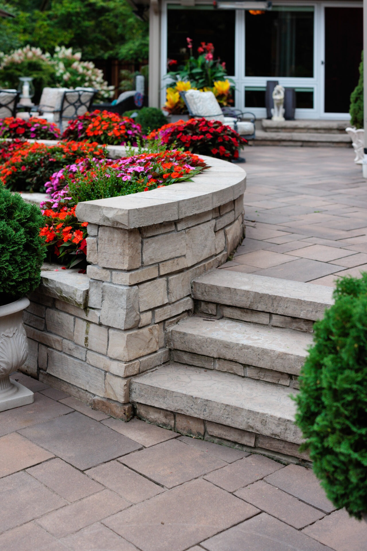 Stone patio with colorful flower garden, steps, and outdoor seating. Well-maintained greenery and decorative elements create a relaxing atmosphere.