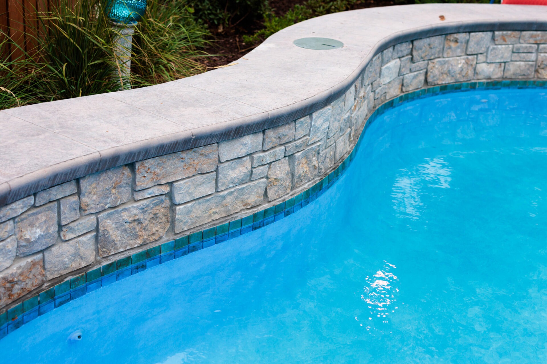 A clear blue swimming pool with a decorative stone edge, surrounded by plants, reflecting sunlight in a tranquil outdoor setting.