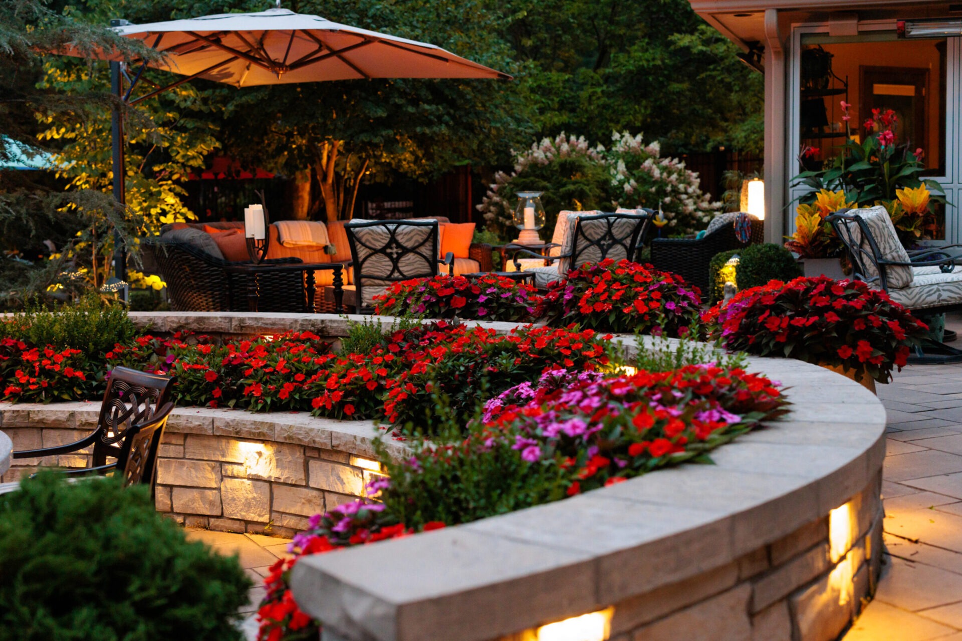 A cozy garden patio with vibrant flowers, elegant seating, warm lighting, and trees, creating an inviting and tranquil outdoor living space.