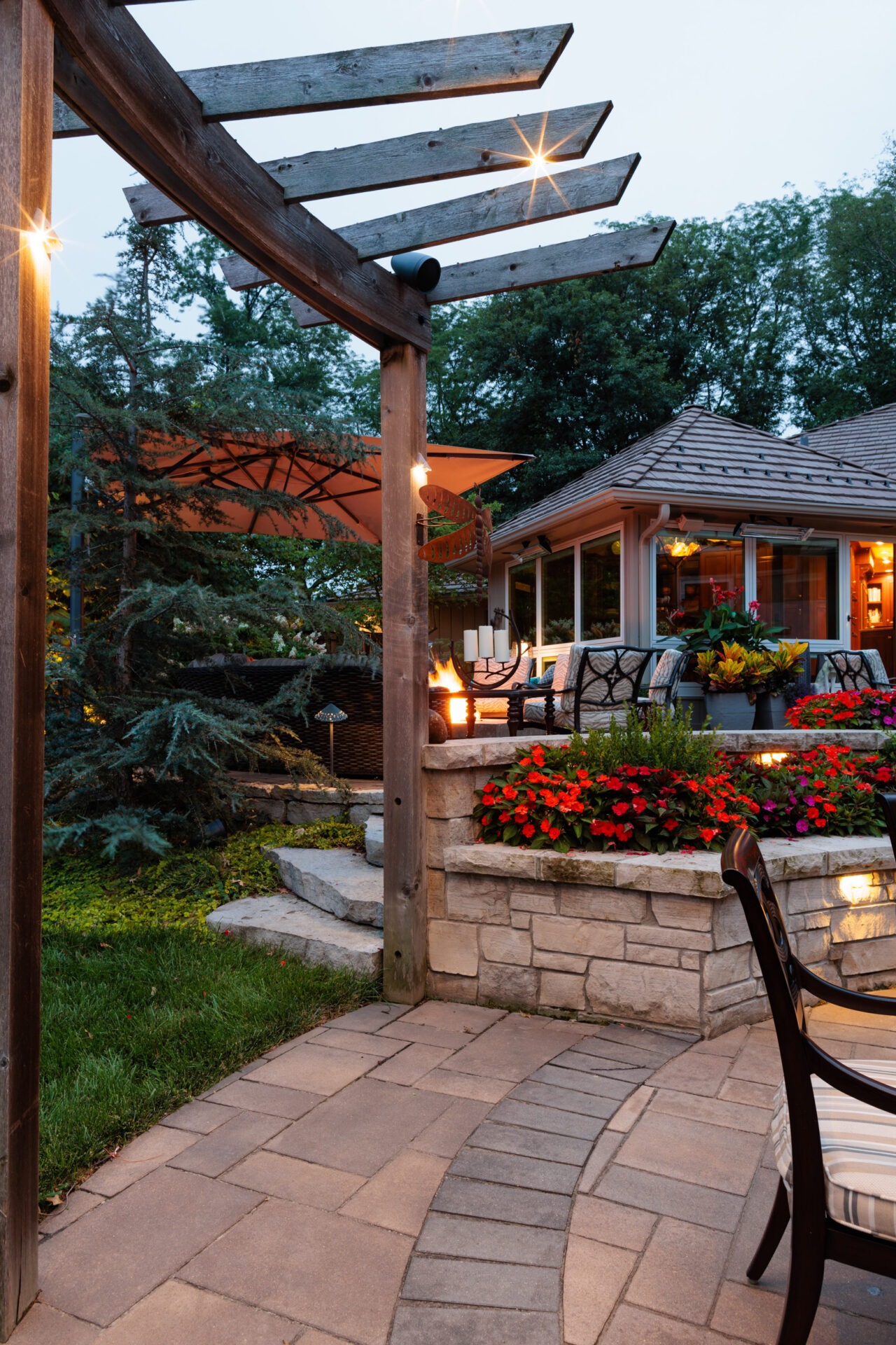 Cozy patio with stone steps, vibrant flowers, pergola, and outdoor seating. Warm lighting creates an inviting atmosphere surrounded by lush greenery.