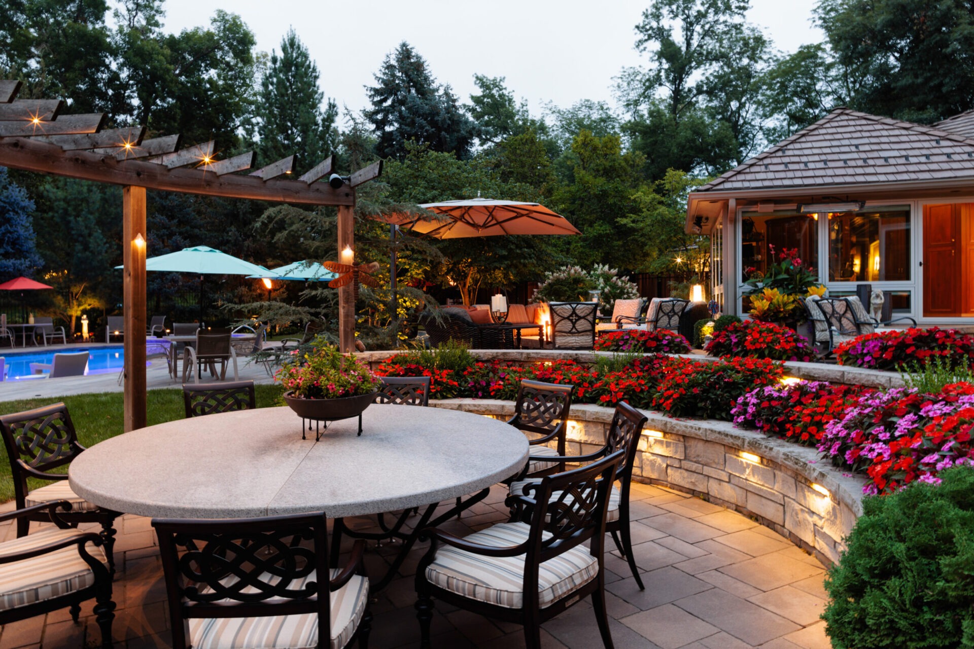 Cozy garden scene with patio furniture, vibrant flowers, a pergola, and a lit pool area surrounded by lush trees and shrubs.