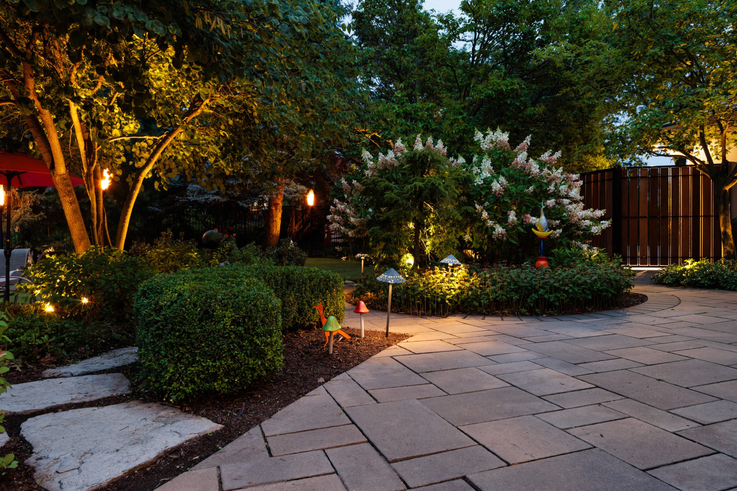 Illuminated garden path at dusk, surrounded by lush greenery and decorative sculptures, creating a serene, enchanting outdoor atmosphere with soft lighting accents.