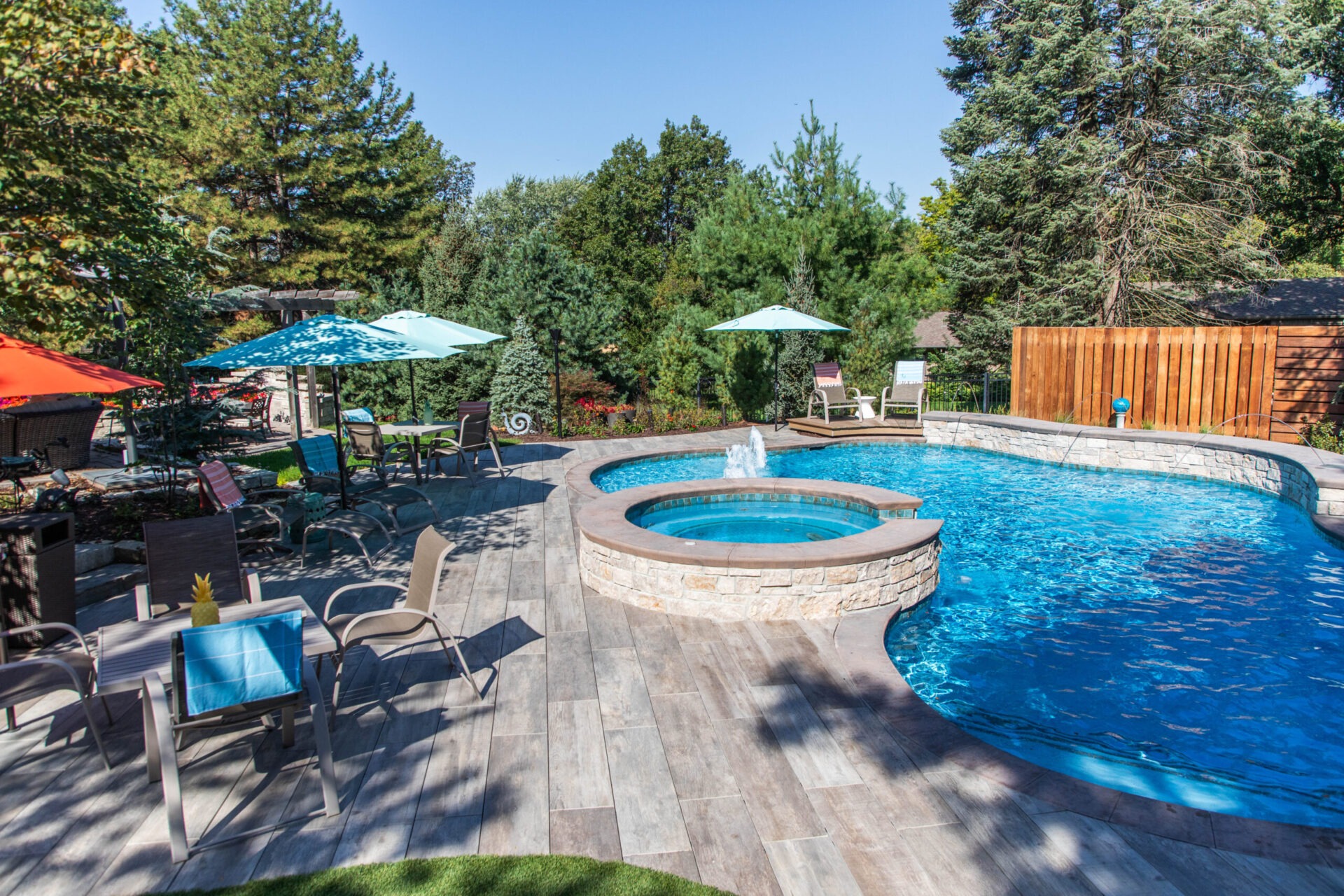 Sunny backyard with a swimming pool and hot tub, surrounded by trees and lounge chairs under colorful umbrellas on a patio.