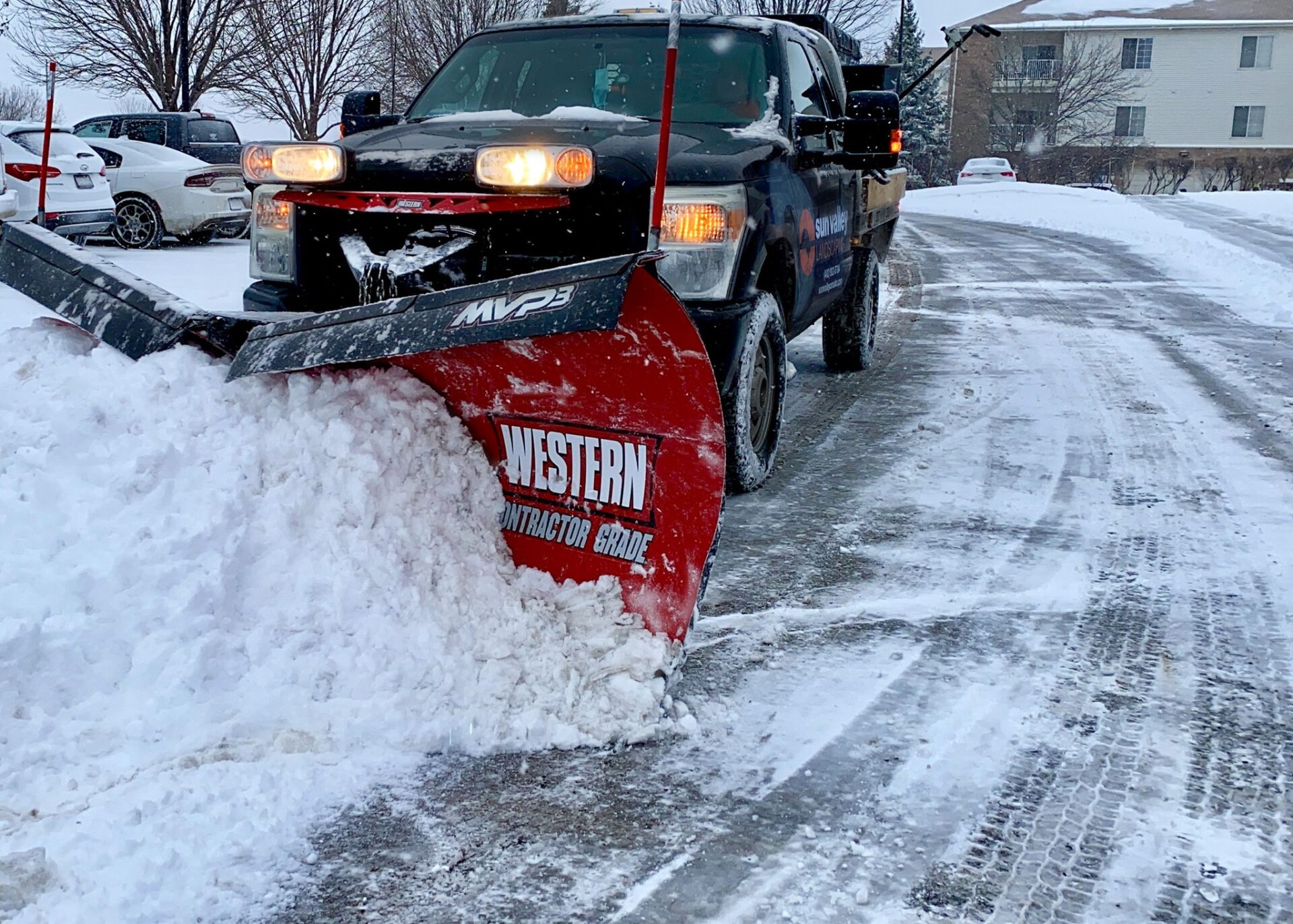 A snow plow truck clearing snow from a parking lot, with cars and apartments in the background, on a cold, snowy day.