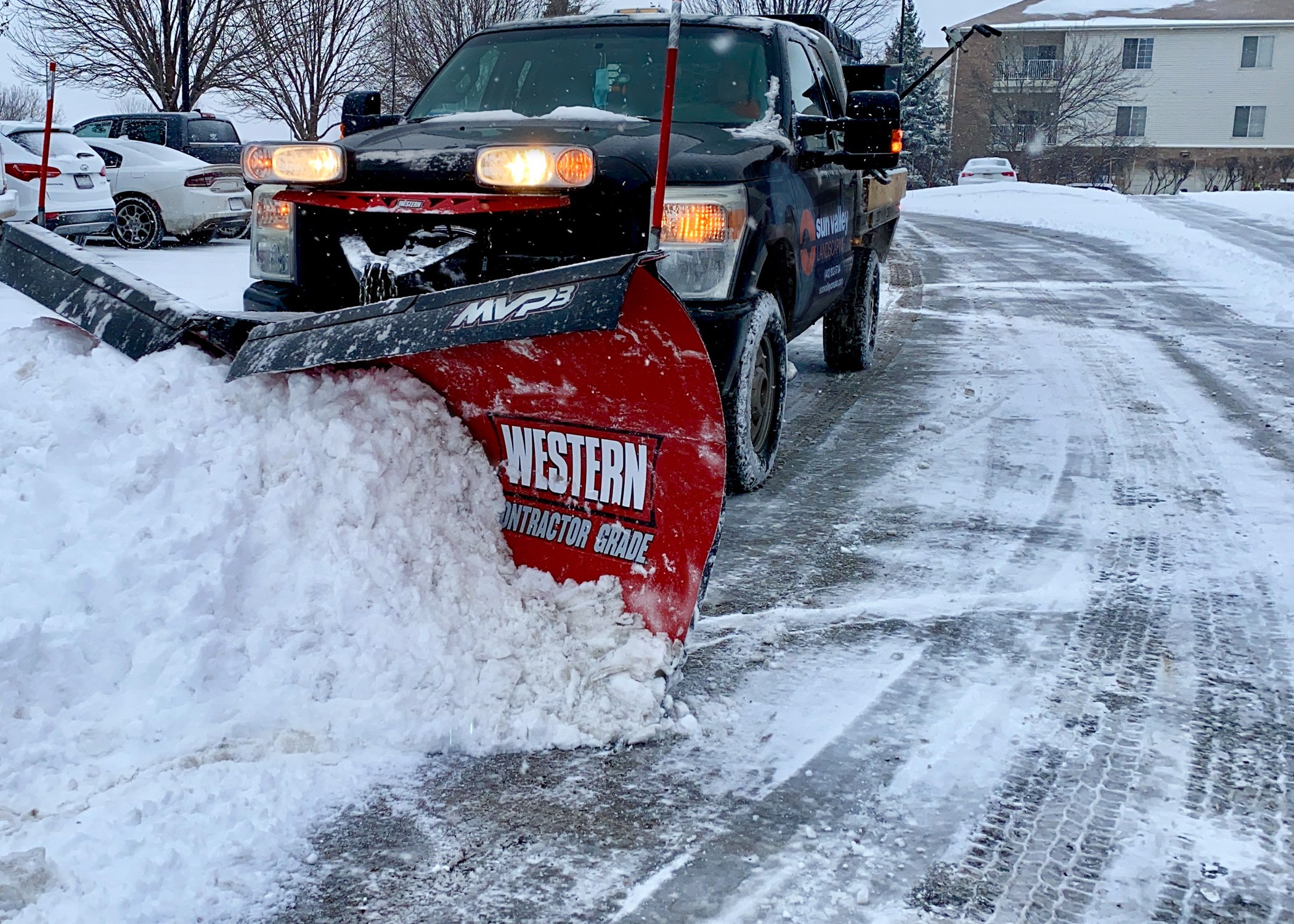 A snowplow truck clearing snow from a residential driveway on a cold winter day, surrounded by parked cars and leafless trees.