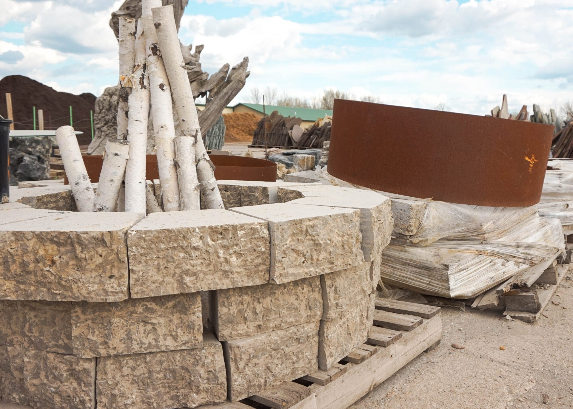 Stone and metal fire pit structure, surrounded by birch logs and pallets, set on sandy ground under a partly cloudy sky.