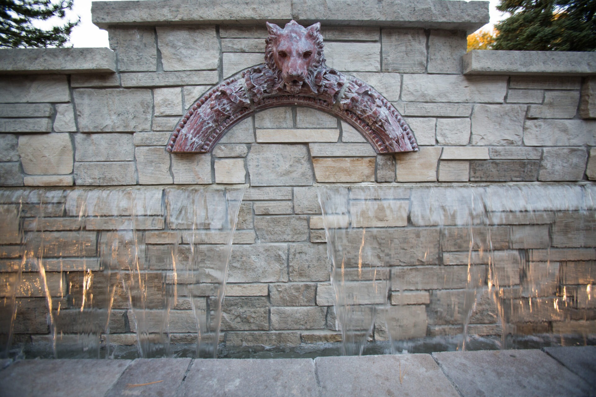 Stone fountain with cascading water, featuring a decorative lion head sculpture. Surrounded by brick wall, creating a calming zen atmosphere.