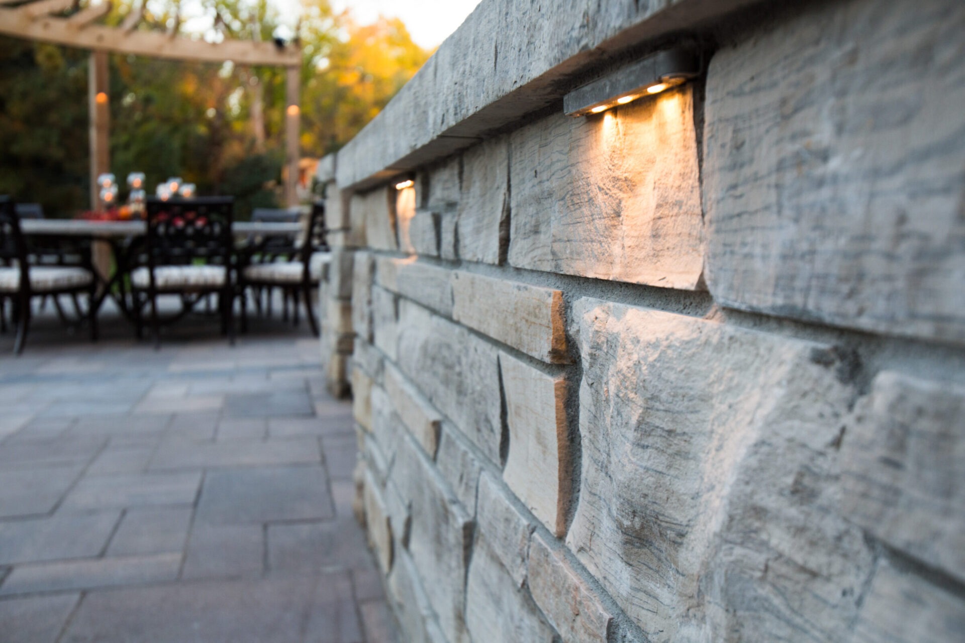 Stone wall with embedded lights lines a patio area. Outdoor dining setup visible in the background under a wooden pergola.