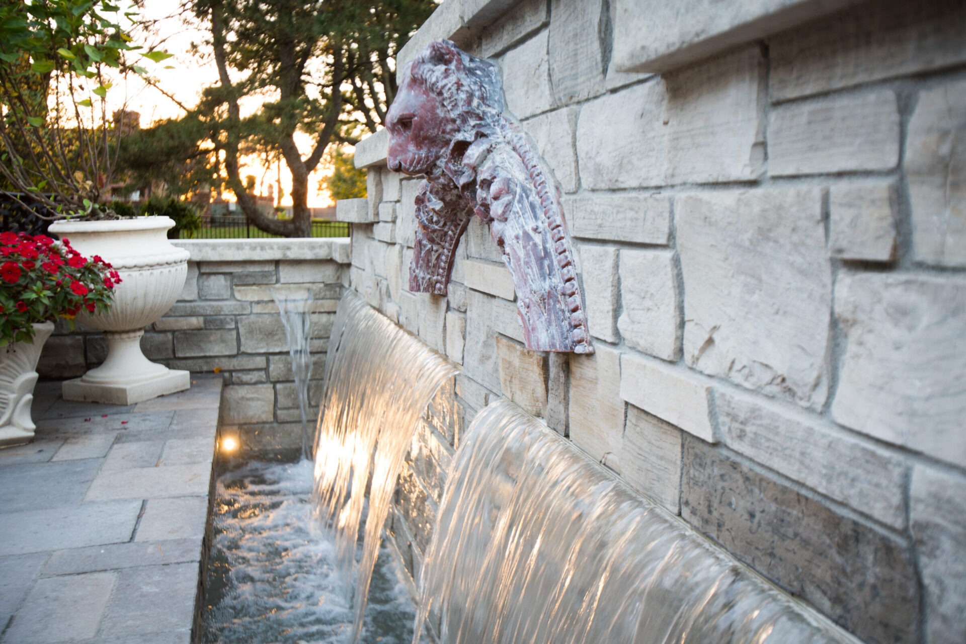 Stone wall fountain with a lion's head sculpture, flowing water, decorative urns with flowers, and trees in the background at sunset.