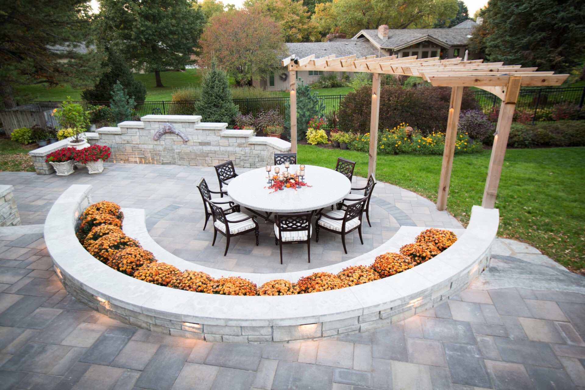 A landscaped garden features a circular stone patio with chairs, table, flowers, and pergola. Greenery and a house are visible.