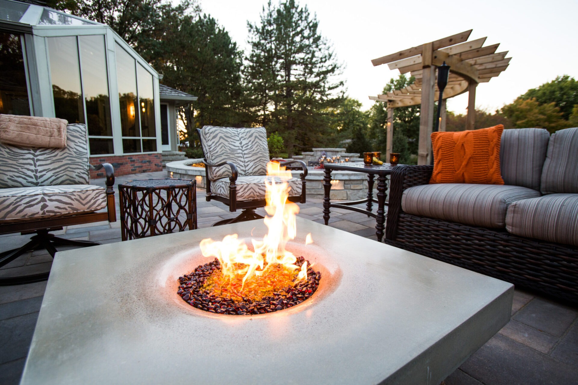 Cozy outdoor patio with cushioned seating surrounds a modern fire pit. Trees and a wooden pergola enhance the serene, inviting atmosphere.