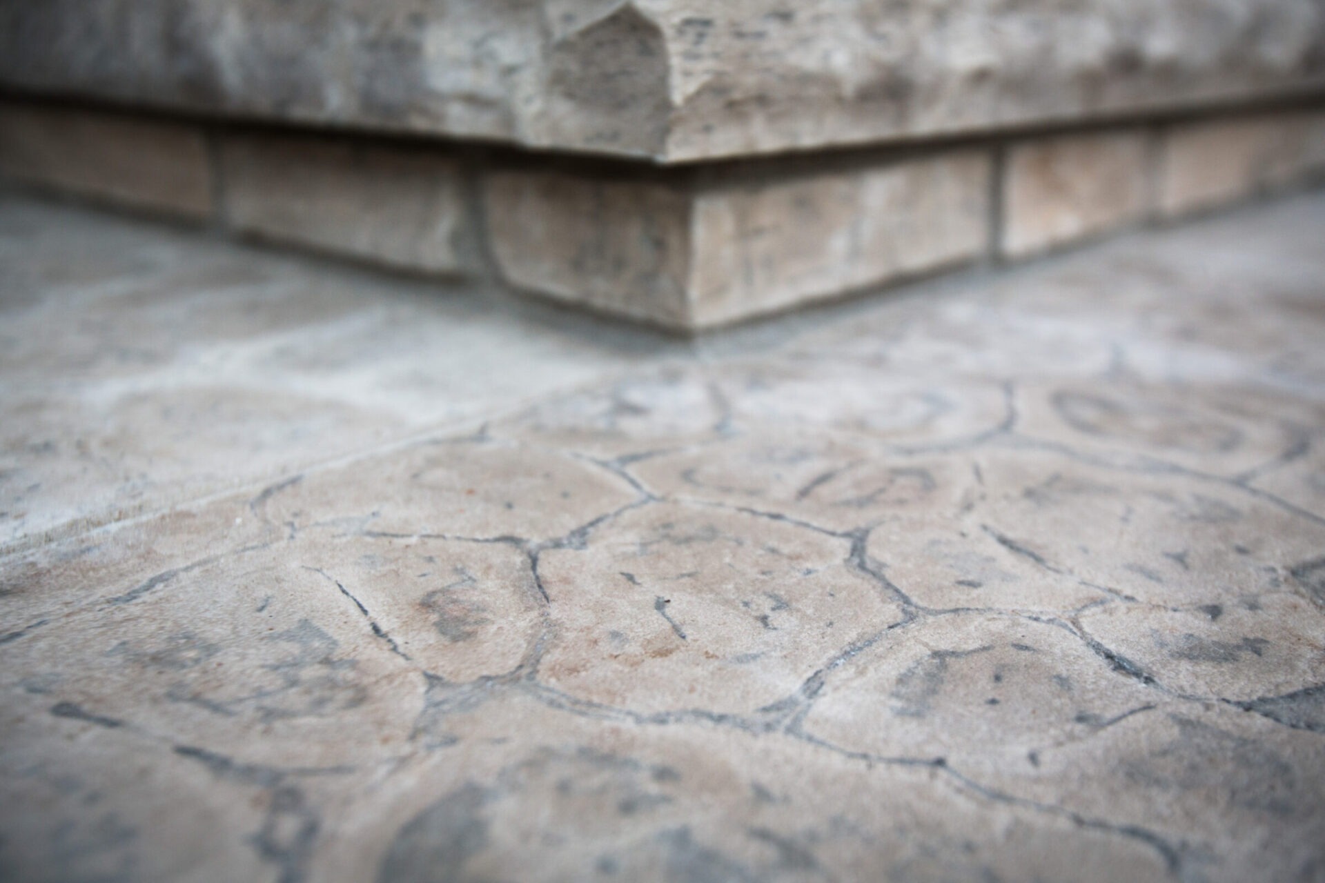 Close-up of textured paving stones and a building foundation corner, showing detailed patterns and natural tones in the stonework, with soft lighting.