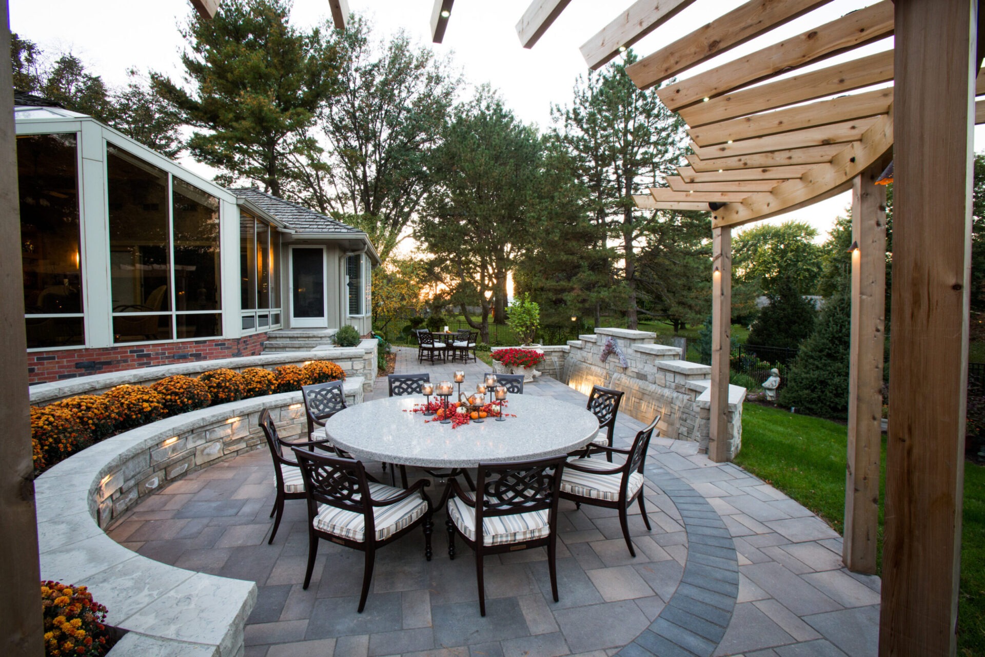 A spacious patio with a round table, surrounded by chairs and flowers, adjacent to a house, amidst lush green trees at sunset.