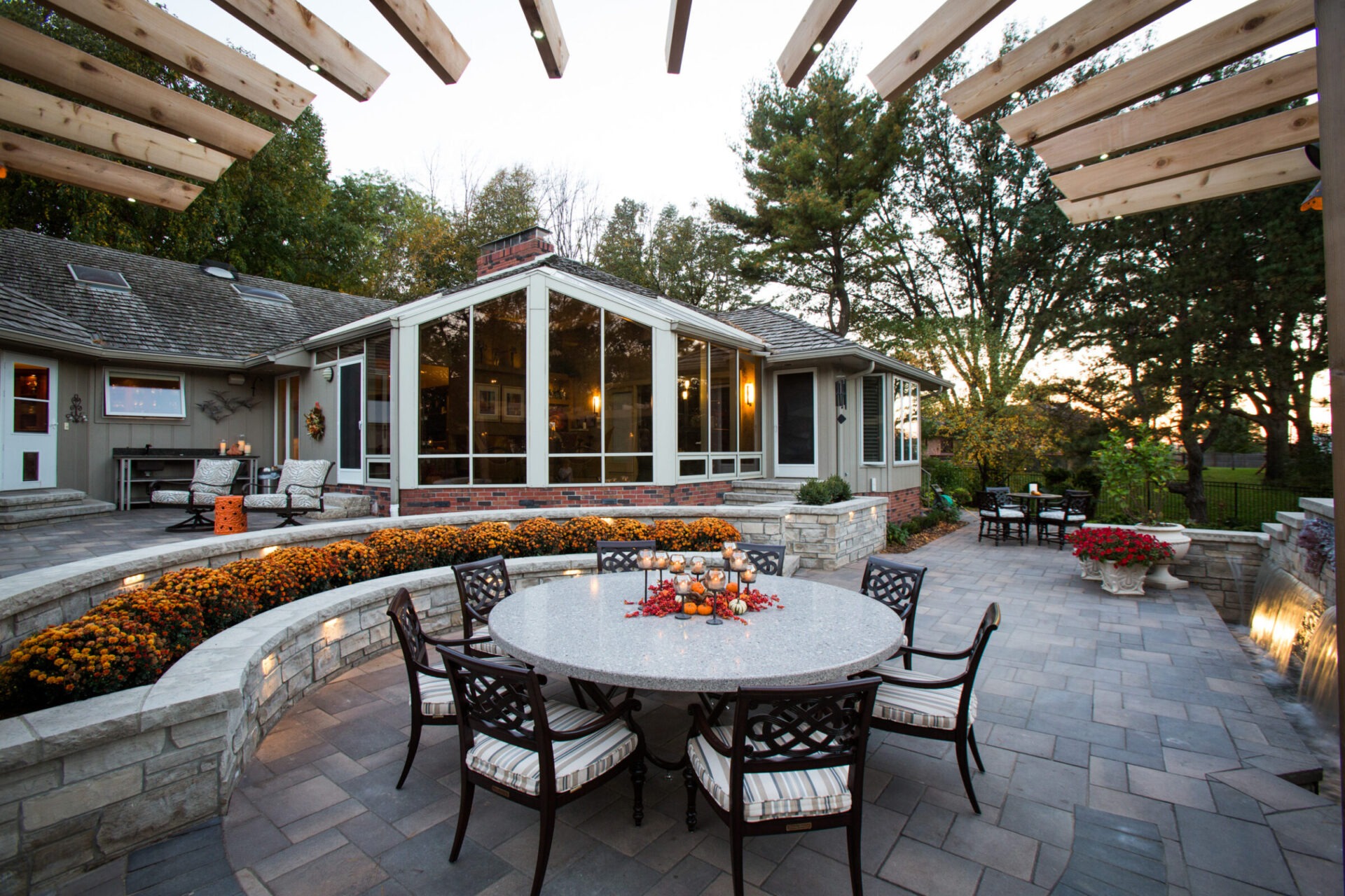 Contemporary backyard with patio furniture, stone flooring, and decorative plants. A modern house features large windows, surrounded by trees and evening ambiance.