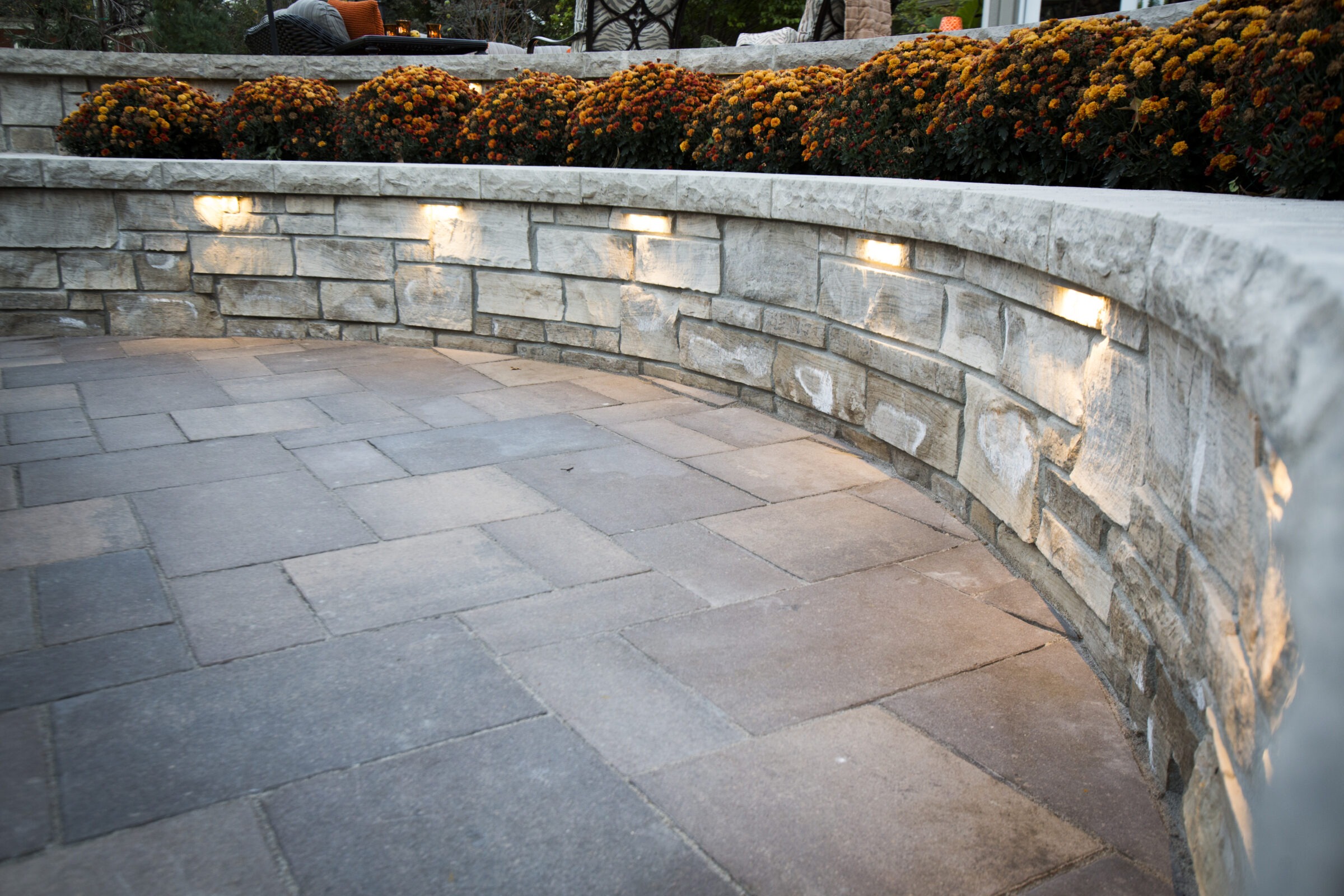 Curved stone wall with embedded lights, bordered by orange flowers. Overlooking a raised patio with outdoor seating and greenery.