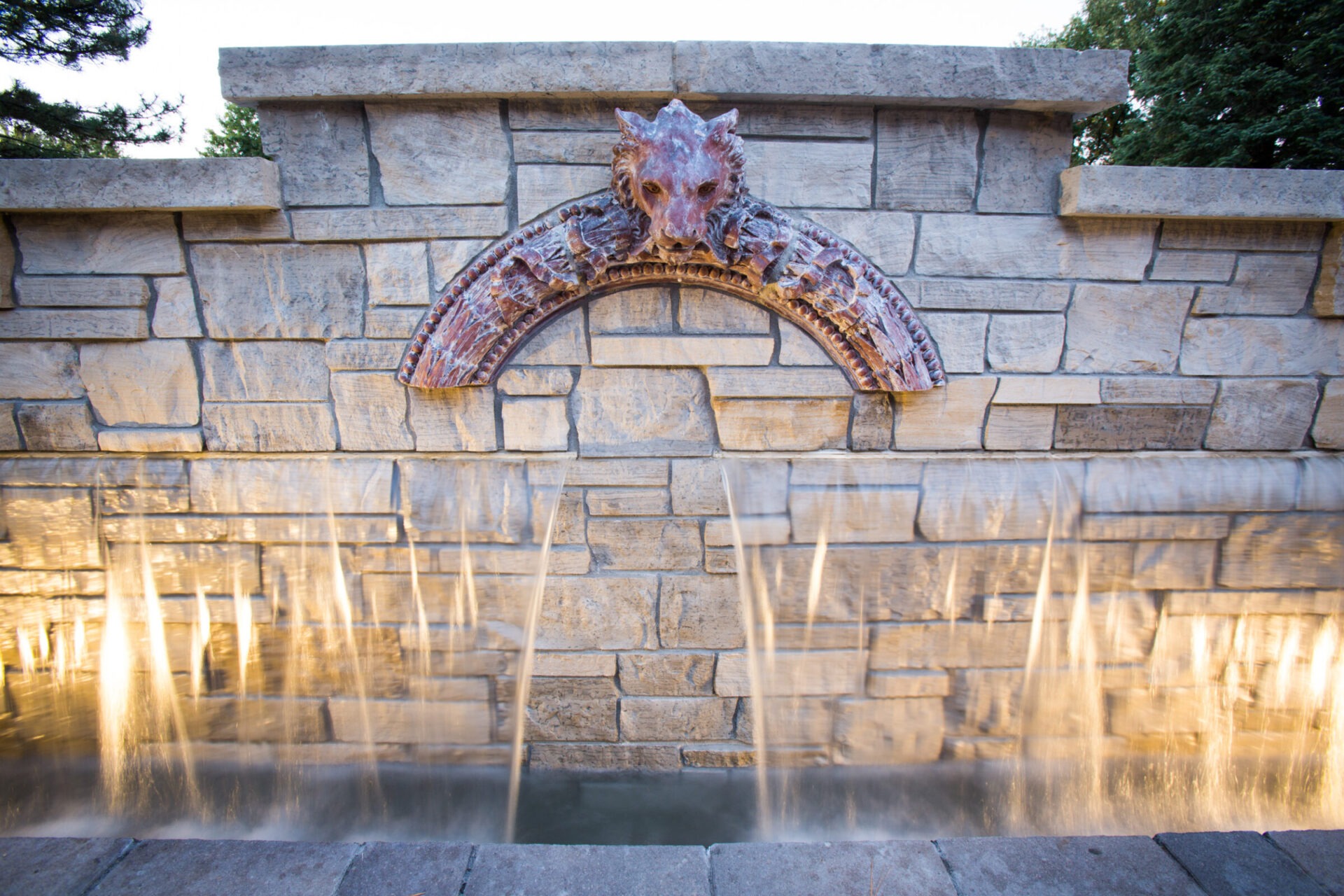 A stone wall with water cascading from three spouts, featuring a central decorative sculpture, surrounded by trees and soft lighting.