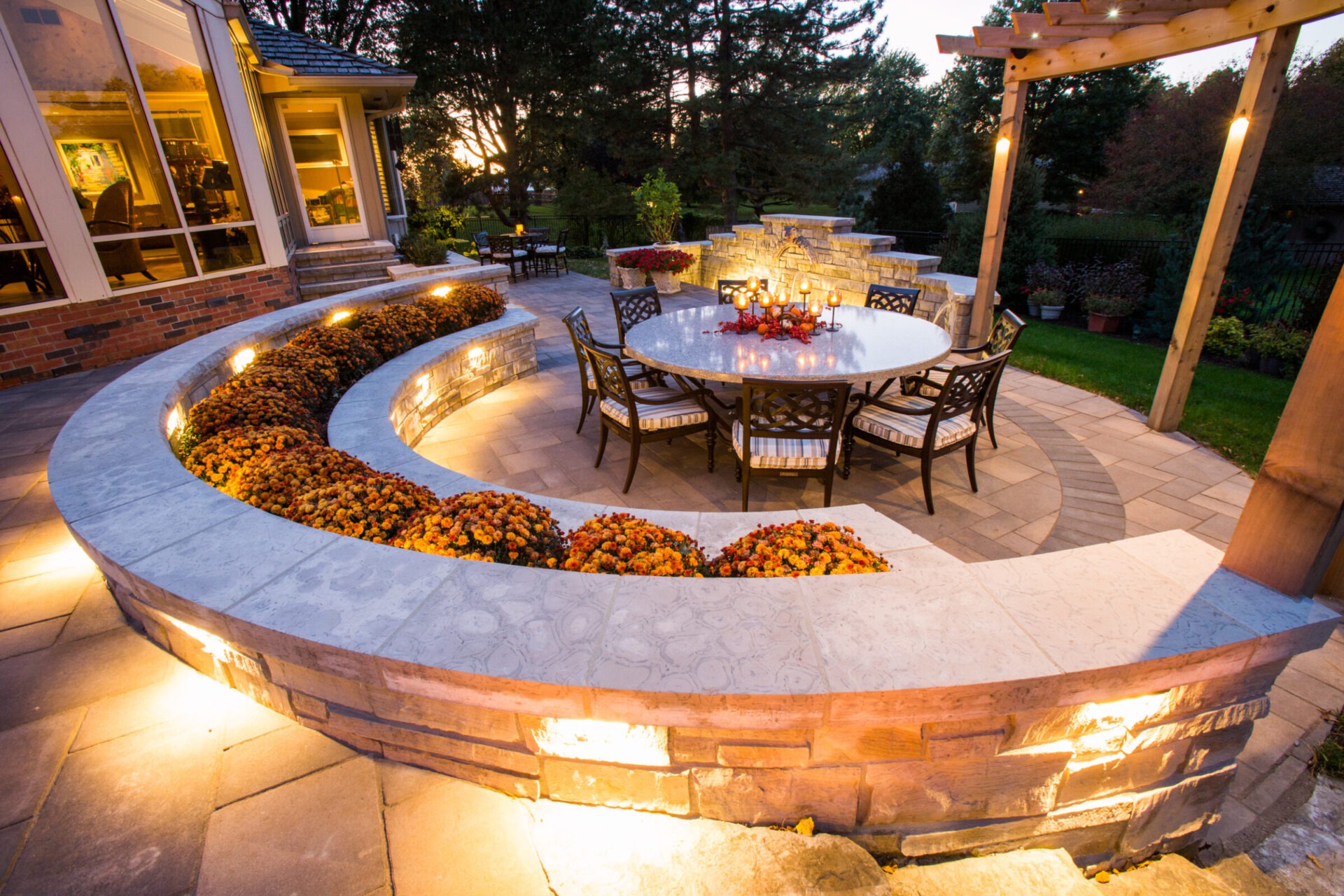 Elegant outdoor patio features curved stone wall, round table with chairs, vibrant flowers, and warm evening lighting, creating a cozy, inviting atmosphere.